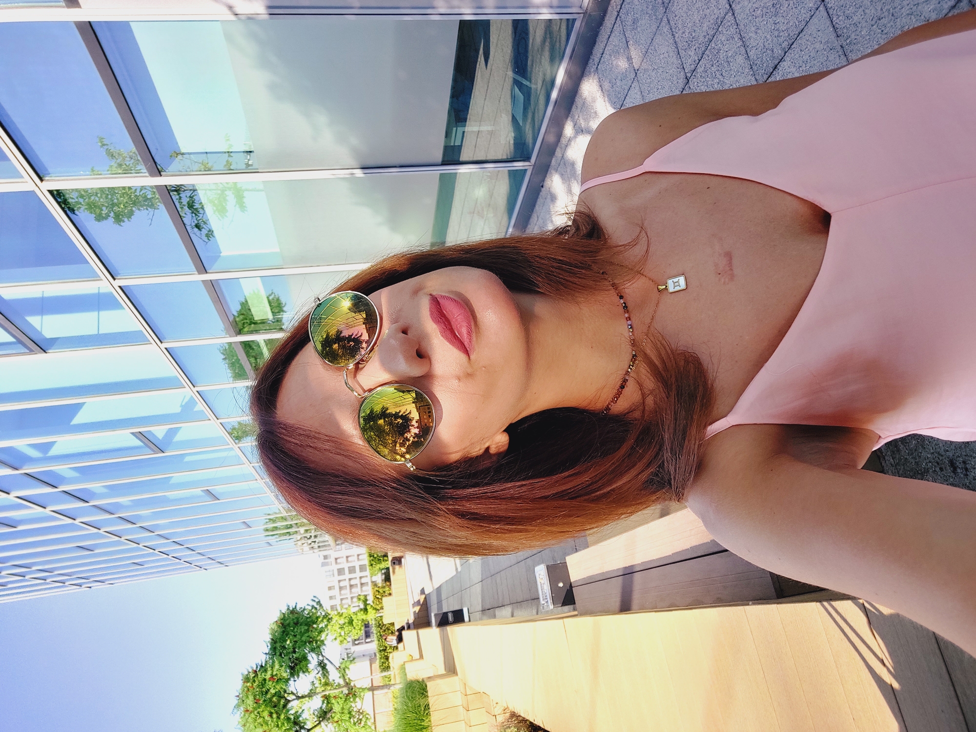 In a close-up, eye-level, outdoor selfie, a woman with reddish-brown hair and light skin is positioned in the center of the frame. She is wearing a light pink slip dress and round, gold-rimmed sunglasses that reflect the surrounding environment. Her lips are slightly parted and painted with a matte pink lipstick, and she is looking upwards, away from the camera. A delicate necklace with colorful beads and a white pendant featuring Roman numerals is visible around her neck. A scar is noticeable on her upper chest.
The background reveals a modern architectural setting with a tall building featuring a glass facade that reflects the sky and trees. To the left, a wooden deck or bench is visible, with a walkway made of stone tiles. Green foliage and trees are interspersed throughout the scene, suggesting an urban park or courtyard. The lighting indicates it is daytime, with sunlight casting shadows.