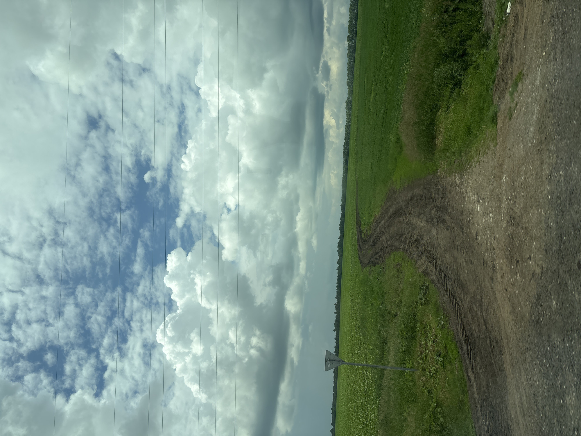 The image depicts a rural landscape with a dirt road winding through green fields under a cloudy sky. The road appears to be unpaved and shows tire tracks. To the left of the road, a triangular road sign on a pole stands in a field of green crops. In the distance, a line of trees forms the horizon. The sky is filled with a mix of white and dark gray clouds, suggesting the possibility of changing weather. The scene is set in the city of Chirsha-Tartysh, Russia. No people or specific actions are visible.
