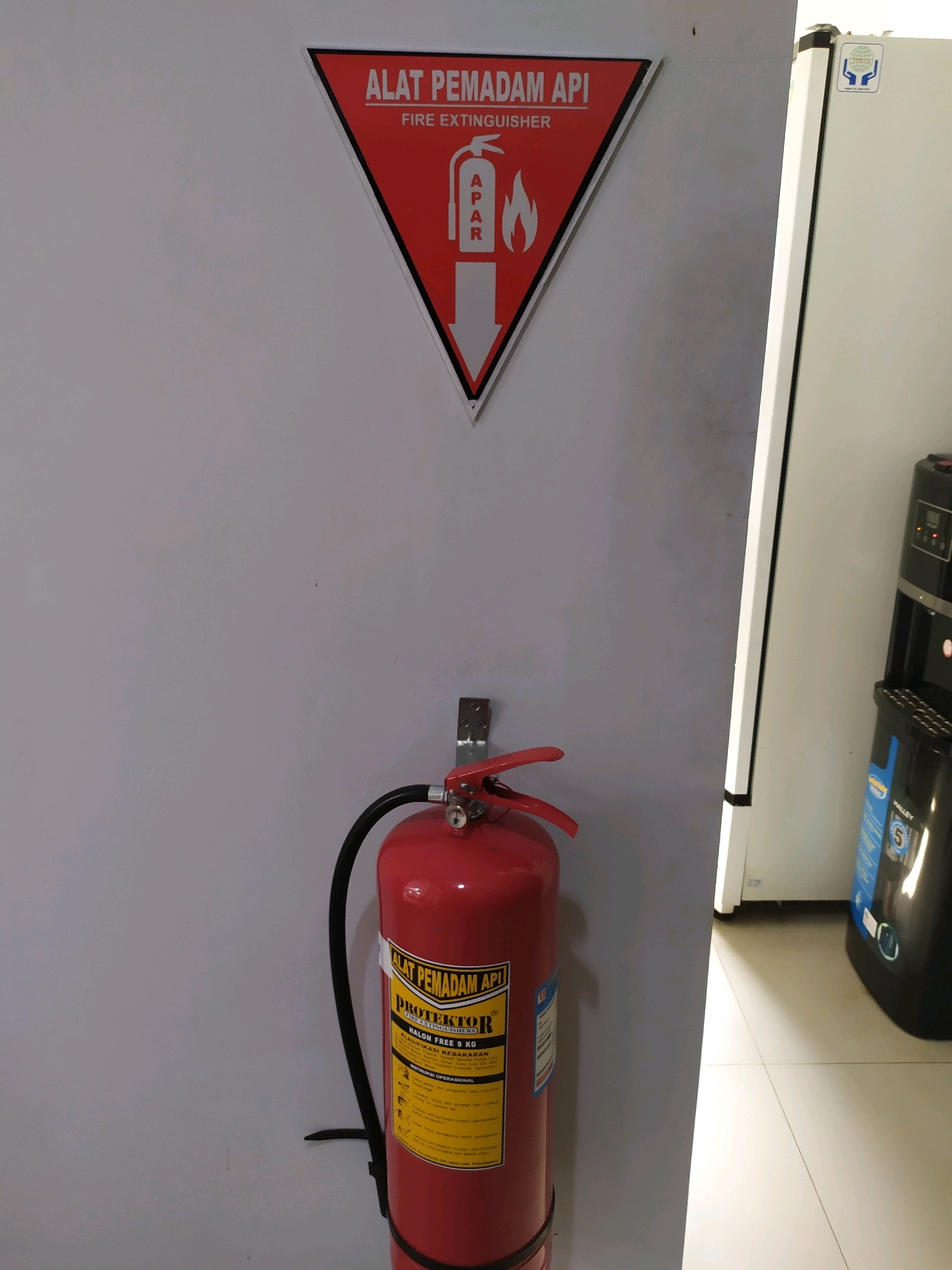 The image shows a fire extinguisher mounted on a pale wall. Above the extinguisher, a red triangular sign with black trim displays "ALAT PEMADAM API" and "FIRE EXTINGUISHER" in white lettering. Inside the triangle, a white graphic depicts a fire extinguisher with the letters "APAR" stacked vertically and a flame symbol. The red fire extinguisher itself has a black hose and a yellow label with text in Indonesian. To the right of the wall and extinguisher, a portion of a white refrigerator and a black water dispenser are visible. The floor appears to be tiled. The scene suggests an indoor environment, likely a public building or facility within the city of Wates, Indonesia.