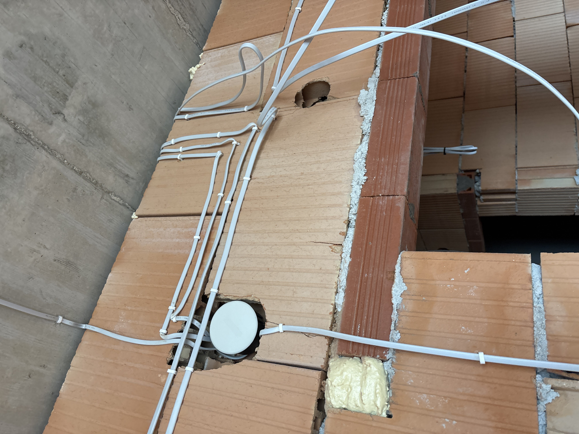 The image shows an interior construction scene, likely within a building. Visible are brick walls, some with a reddish-brown hue, constructed with mortar or expanding foam. White electrical conduits are attached to the brick walls with plastic clips. The conduits appear to be wired for electrical or communication purposes. A round, white electrical box is visible, embedded in one of the brick walls. A concrete ceiling is partially visible in the upper left of the frame.
There are no people visible in the image, nor is there sufficient information to determine if it is the city of Burgas, Bulgaria.