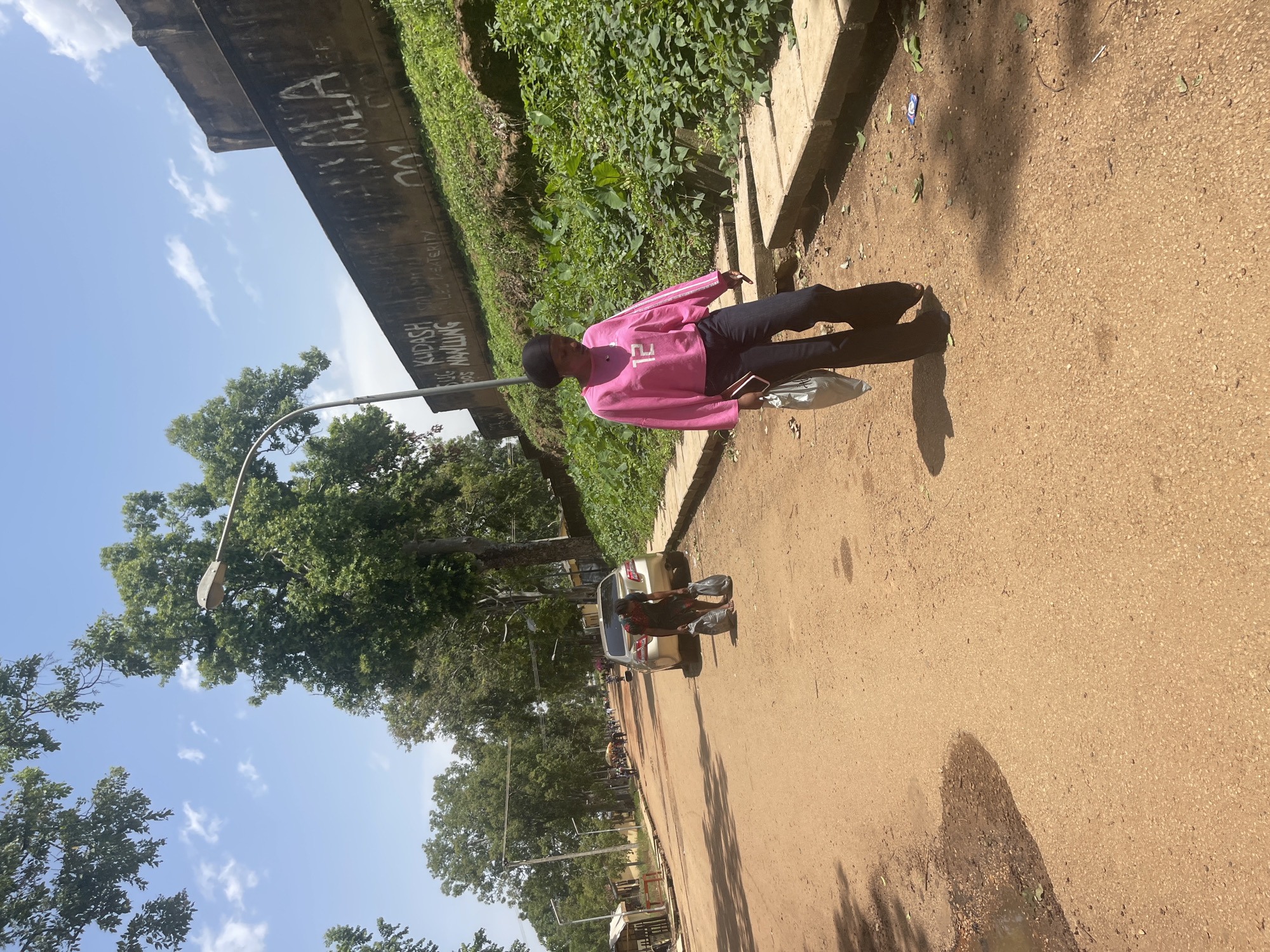 The image, taken in Rafin Par, Nigeria, depicts a person standing on a dirt road. The individual is wearing a pink long-sleeved shirt with white stripes on the sleeves and black pants. They are holding a metallic-colored bag or purse in their right hand and a phone in their left. The person is facing away from the camera, with their head turned to the side, and appears to be walking.
The setting is outdoors, with a dusty, light brown dirt road occupying the foreground and middle ground. To the left of the road, a concrete barrier runs along the edge, behind which is a steep embankment covered in lush green foliage. A concrete structure, possibly an overpass or bridge, is visible in the upper left portion of the image, with graffiti on its underside. Trees with green leaves line the road further down, and a street lamp is present.
In the background, a silver car is parked on the side of the road, with another person, partially obscured, sitting in the driver's seat. More vehicles and people are visible further down the road, suggesting a somewhat busy street or thoroughfare.
The sky is blue with some scattered white clouds, indicating a bright, possibly sunny day. The sun casts shadows on the road, suggesting it is daytime, likely late morning or afternoon. The overall mood is casual and everyday, capturing a moment in a common outdoor setting.
Visible text includes graffiti on the underside of the concrete structure, though it is partially obscured and difficult to read clearly. Some phrases like "KUDASH" and "NAWLING" are discernible.