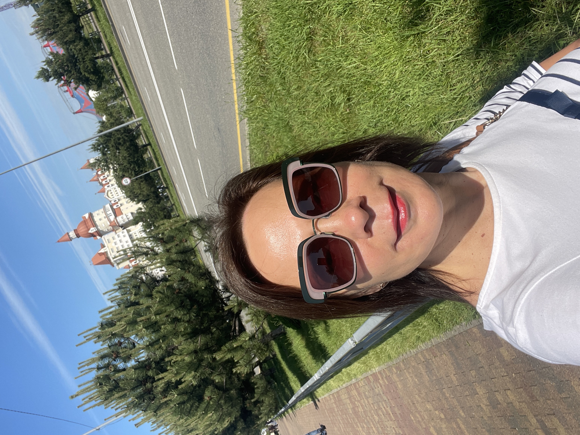 A woman is lying on the grass in an outdoor setting. She is wearing sunglasses and a white T-shirt with stripes. In the background, there is a road with white lines, grass, trees, and a building with red towers. The building appears to be located in the city of Sirius, Russia, as evidenced by its architectural style, consistent with that found in the city.