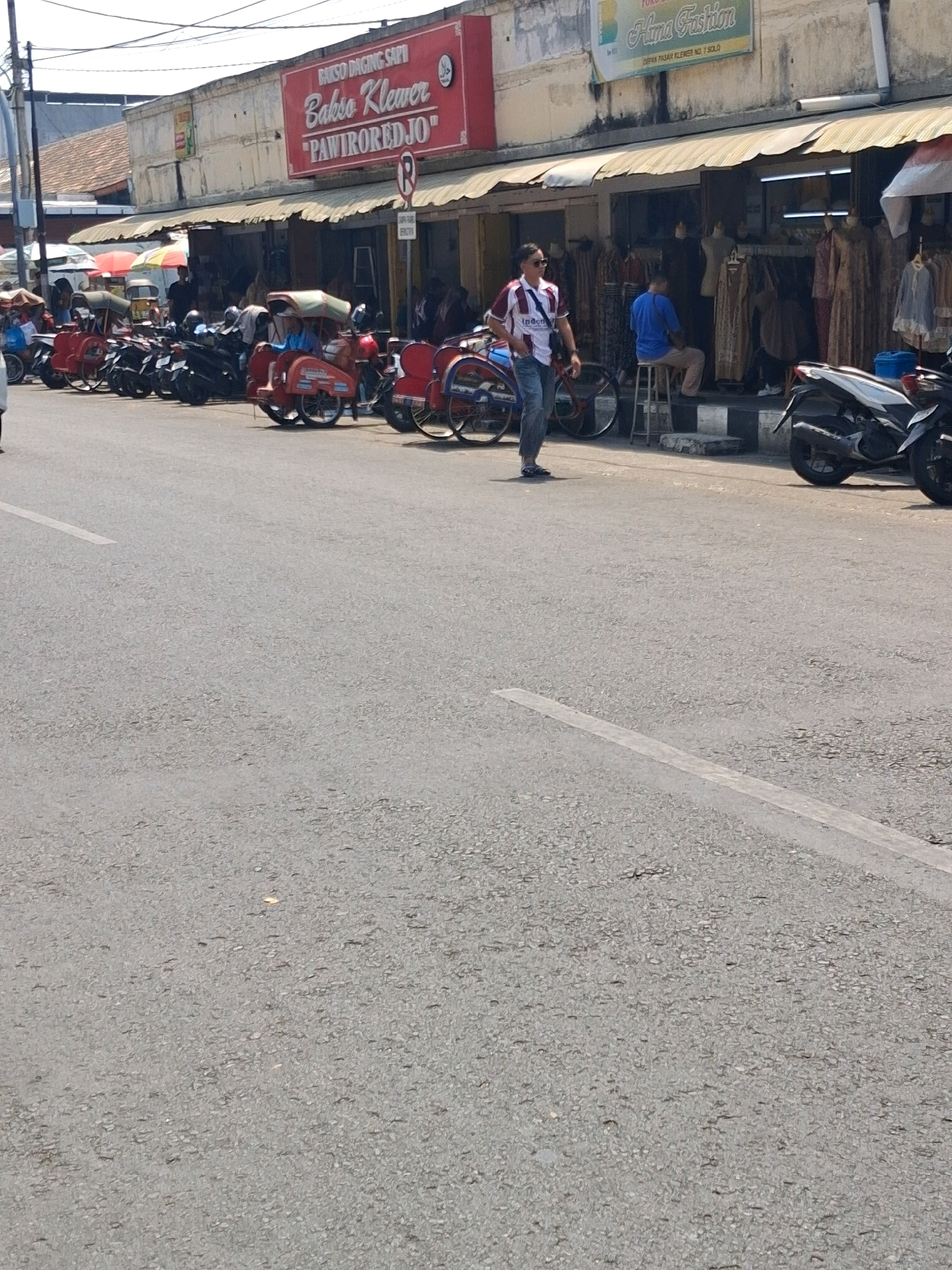 The image depicts a street scene in Surakarta, Indonesia, likely in the vicinity of Klewer Market. In the background, a row of shops lines the street, with awnings providing shade. One shop prominently displays a red sign that reads "BAKSO DAGING SAPI Bakso Klewer 'PAWIROREDJO'". Another shop has a green sign that says "Hima Fashion". The street is lined with parked motorcycles and several becaks (pedicabs), some of which are occupied by people. A man wearing a white and red striped shirt and jeans is walking on the sidewalk. Another man in a blue shirt is sitting on a stool in front of a shop. The foreground is dominated by an asphalt road with a white lane marking. The environment appears to be a busy commercial area with a mix of traffic and pedestrian activity. The overall lighting suggests it is daytime, with bright sunlight casting shadows.