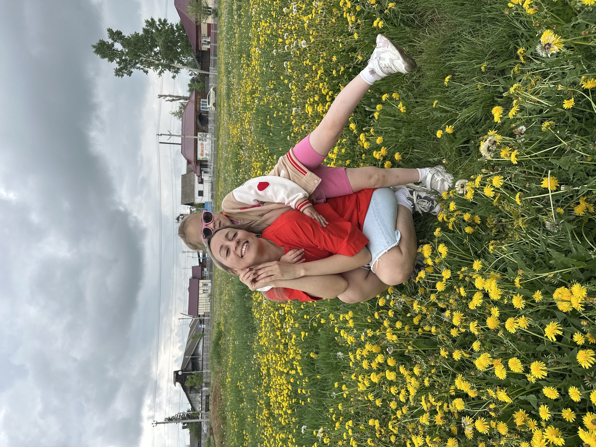 A woman and a young girl are pictured crouching in a field of yellow dandelions. The woman is wearing a red t-shirt and blue denim shorts, while the girl is wearing pink shorts and a beige and white jacket with red trim and a heart detail. The girl is embracing the woman from behind, and both are smiling. In the background, there are houses, a power line pole, a tall tree, and a cloudy sky. The overall scene suggests a casual outdoor activity in a rural or suburban setting, likely in Dzerzhinskoye, Russia.