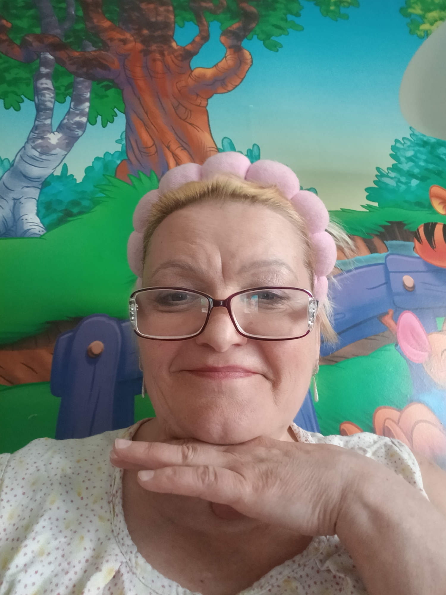 A woman with blonde hair and glasses is posing for a selfie in front of a wall mural depicting a cartoon forest. She is wearing a pink headband and a white top with yellow polka dots. Her left hand is resting under her chin. Based on the cartoon mural, the location may be a child's room or a childcare facility. There is no evidence to confirm the location is in the city of Красный Маяк, Russia.
