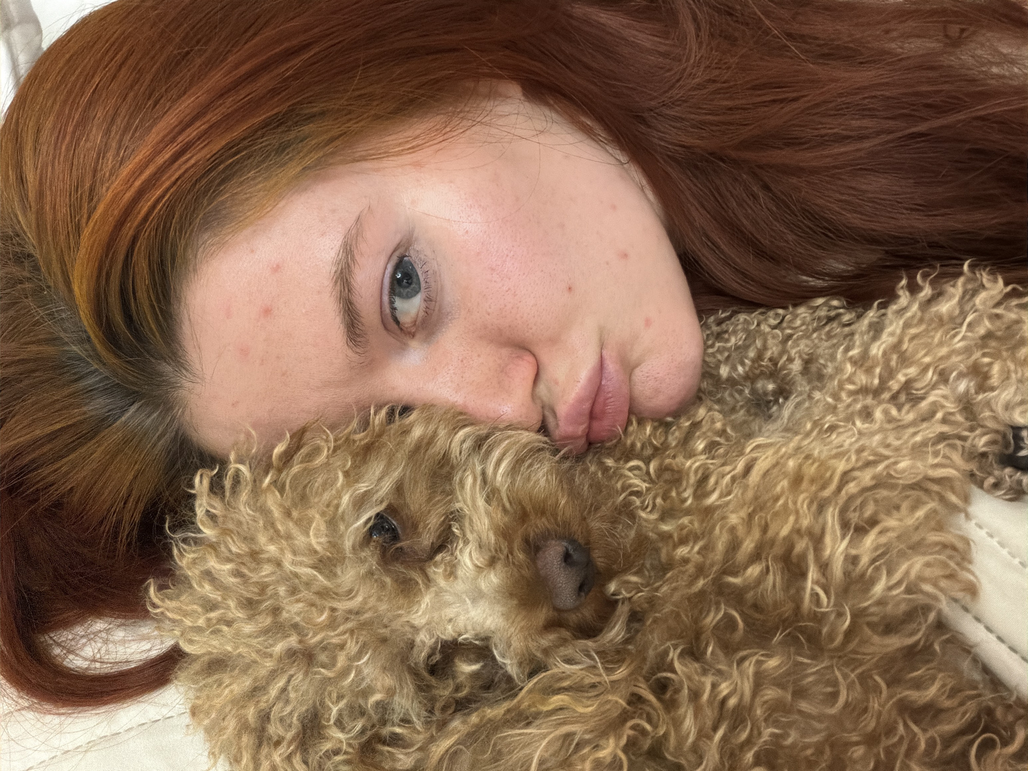 The image shows a close-up, high-angle, medium shot of a young woman with long, reddish-brown hair and a light-skinned complexion, with freckles and some acne visible on her face. She is pouting her lips and appears to be pressing her cheek against the fur of a light brown, curly-haired dog. The dog's eye is visible. The background consists of a textured surface, possibly bedding. The scene appears to be indoors. There is no information provided to indicate the location within Krasnodar, Russia.