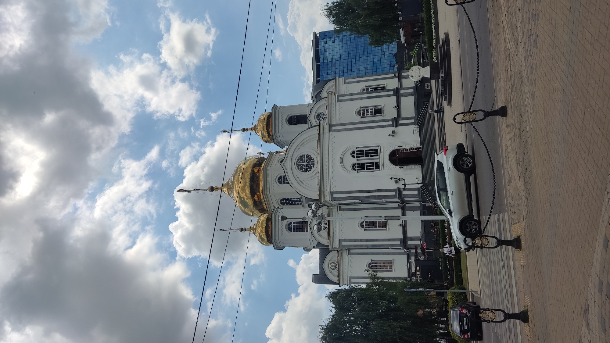 The image shows a white cathedral with golden domes, situated in the city of Krasnodar, Russia. The cathedral is an imposing structure with a classical architectural style, featuring multiple domes and arched windows. In the foreground, a white SUV is parked on the street, with a pedestrian crossing visible. A black car is seen in the distance, on the left side of the road. The sky is partly cloudy, with a mix of sun and shade, indicating a bright day. A modern glass-fronted building is visible in the background, to the right of the cathedral, suggesting an urban setting. The scene appears to be a public space, possibly a plaza or a square, with trees lining the sides and a decorative fence separating the pedestrian walkway from the street. The overall atmosphere is serene and orderly, capturing a moment of everyday life in the city.