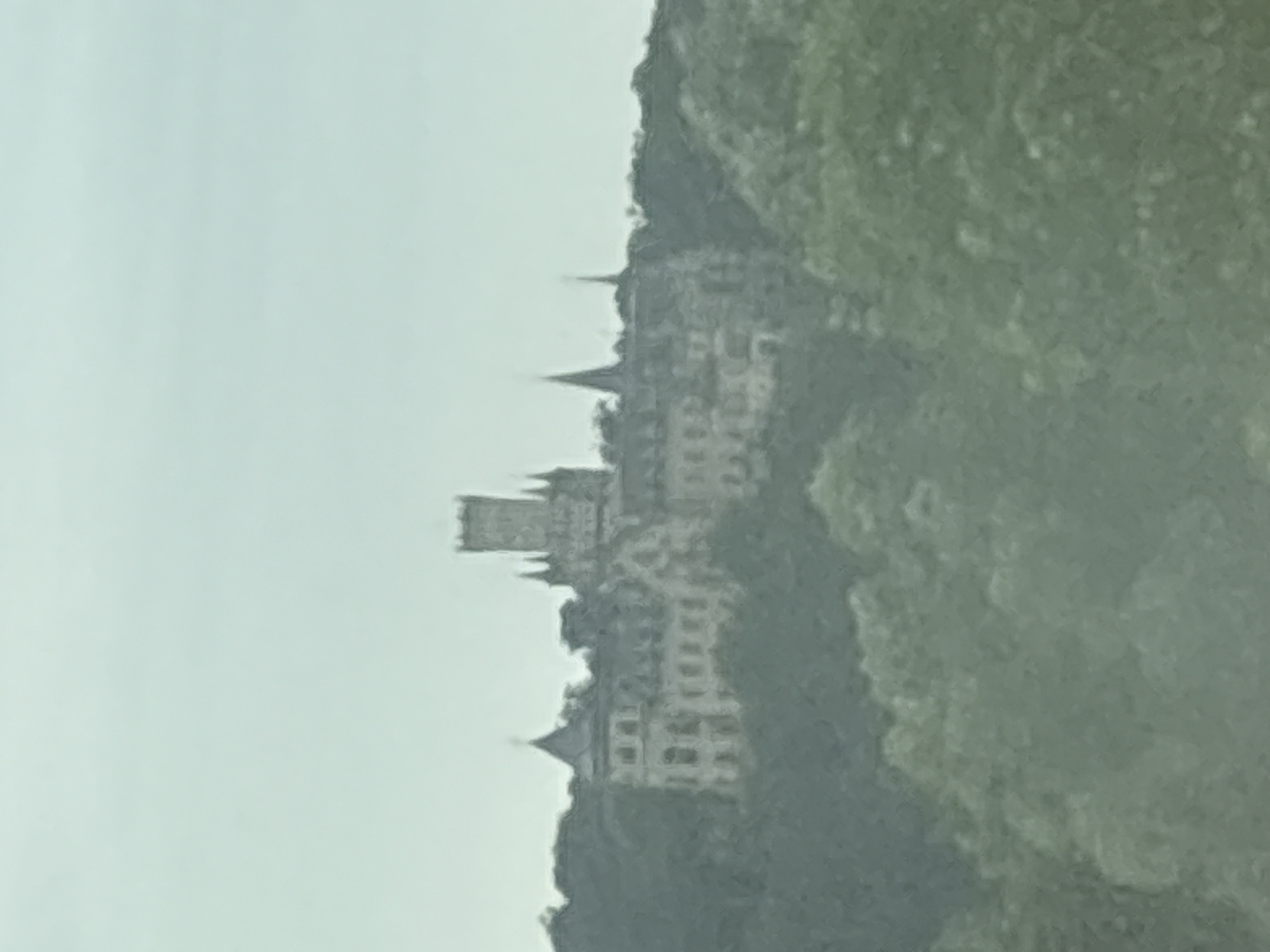 The image shows a pale, overcast sky above a hill covered in trees. Perched on the hill is a large, ornate building that appears to be a castle or a grand manor. The architecture is varied, with multiple turrets and gabled roofs. One prominent feature is a tall, square tower with smaller turrets. The building is light-colored, contrasting with the dark green foliage surrounding it. The overall scene is distant and somewhat hazy, suggesting it was photographed from a considerable distance or through some atmospheric obstruction. This building is Marienburg Castle, located in Nordstemmen, Germany.