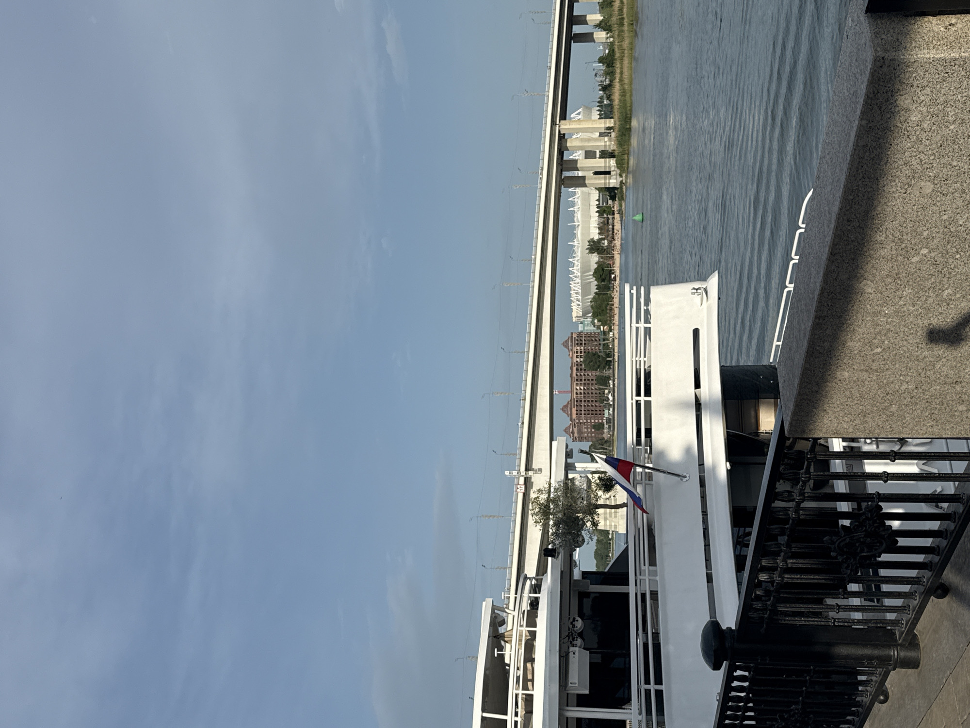 The image depicts a view from the embankment in Rostov-on-Don, Russia. A white boat with the Russian flag is docked along the embankment, with black decorative railings in the foreground. In the background, across the river, is the Rostov Arena stadium. A bridge spans the river, connecting the two sides. The sky is blue with some clouds. The river has a slightly rippled surface. A section of the embankment itself is visible, with a concrete wall.