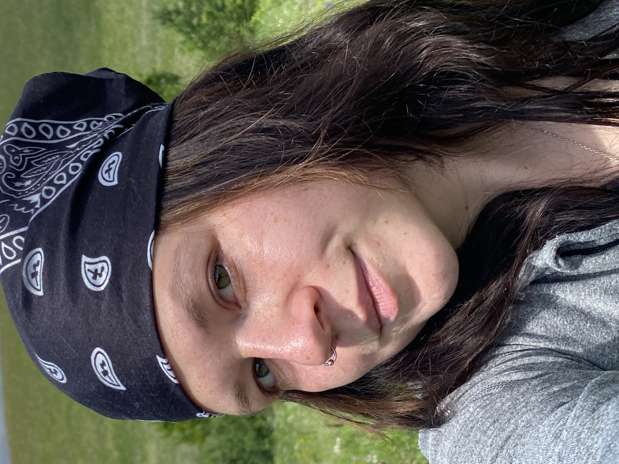 A woman with dark hair, wearing a grey shirt and a black bandana with white paisley patterns, smiles at the camera. She has a nose ring. The background is a blurry green field with trees. The lighting suggests it is daytime. There is no indication that this scene is located in Bogoslovka, Russia.