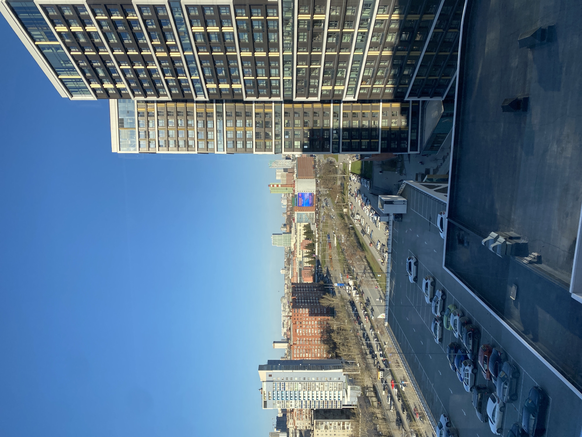 The image shows a high-angle view in Yekaterinburg, Russia. The skyline is punctuated by modern high-rise buildings, one a prominent structure with a grid-like facade reflecting the blue sky. Below, a wide avenue is visible, lined with trees, traffic, and a row of parked cars. Other buildings of varying heights and architectural styles can be seen in the distance. The sky is clear and blue. Adjacent to the parking area, a rooftop surface is visible.