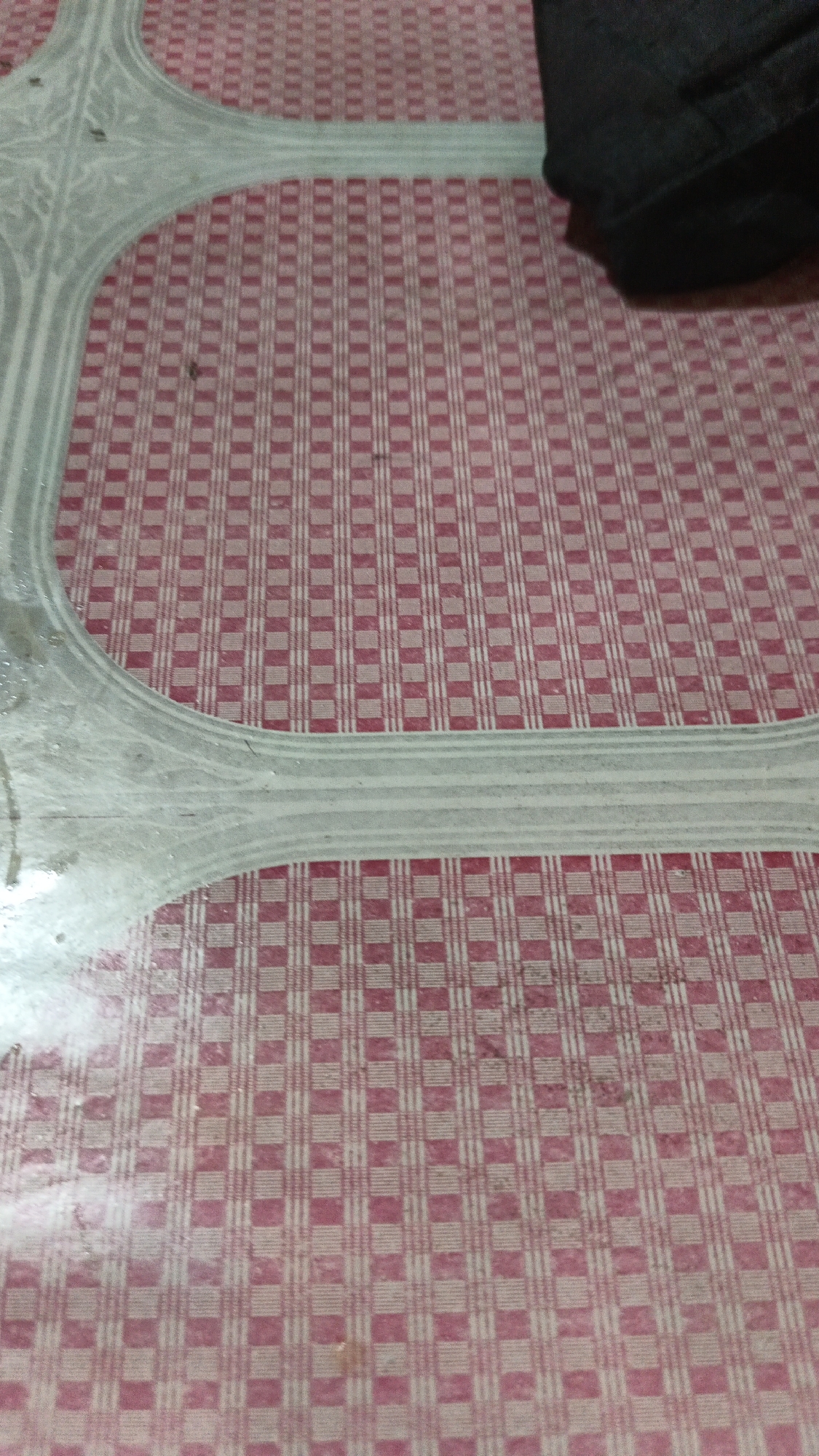 The image shows a close-up, overhead view of a red and white checkered tablecloth. A large, light-colored, decorative border with ornate patterns runs across the left side of the frame, partially obscuring the tablecloth. In the upper right corner, a dark object, possibly a piece of clothing or fabric, is visible. The scene appears to be indoors, given the checkered tablecloth which is a common household item. There is no information in the image to suggest this is located in Dawe, Indonesia, or any other specific city.