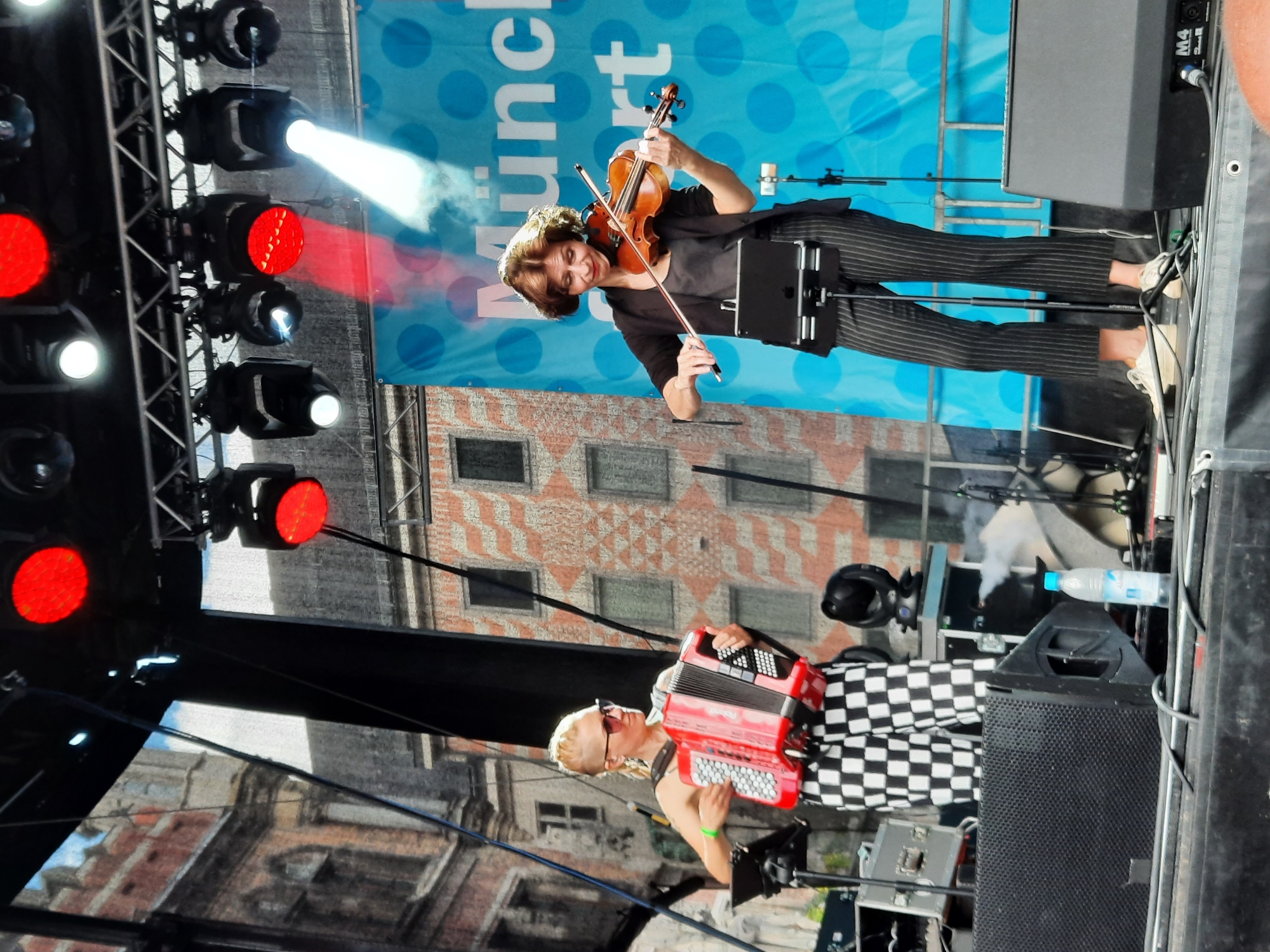 Two women are performing on an outdoor stage. The woman on the left, wearing sunglasses and a blonde mohawk, plays a red accordion. She is dressed in black and white checkered pants. The woman on the right, with short brown hair, plays a violin. She is wearing a black blazer over a dark top and pinstripe pants. Both women are facing forward, towards an unseen audience. Behind them, a blue banner with white polka dots and white text, possibly from "München," is visible. Stage lights are mounted overhead, with some emitting red and white beams. The background includes buildings with red brick and patterned facades. Various audio equipment, including speakers and microphones, are present on the stage. A bottle of water sits on a speaker in the foreground.