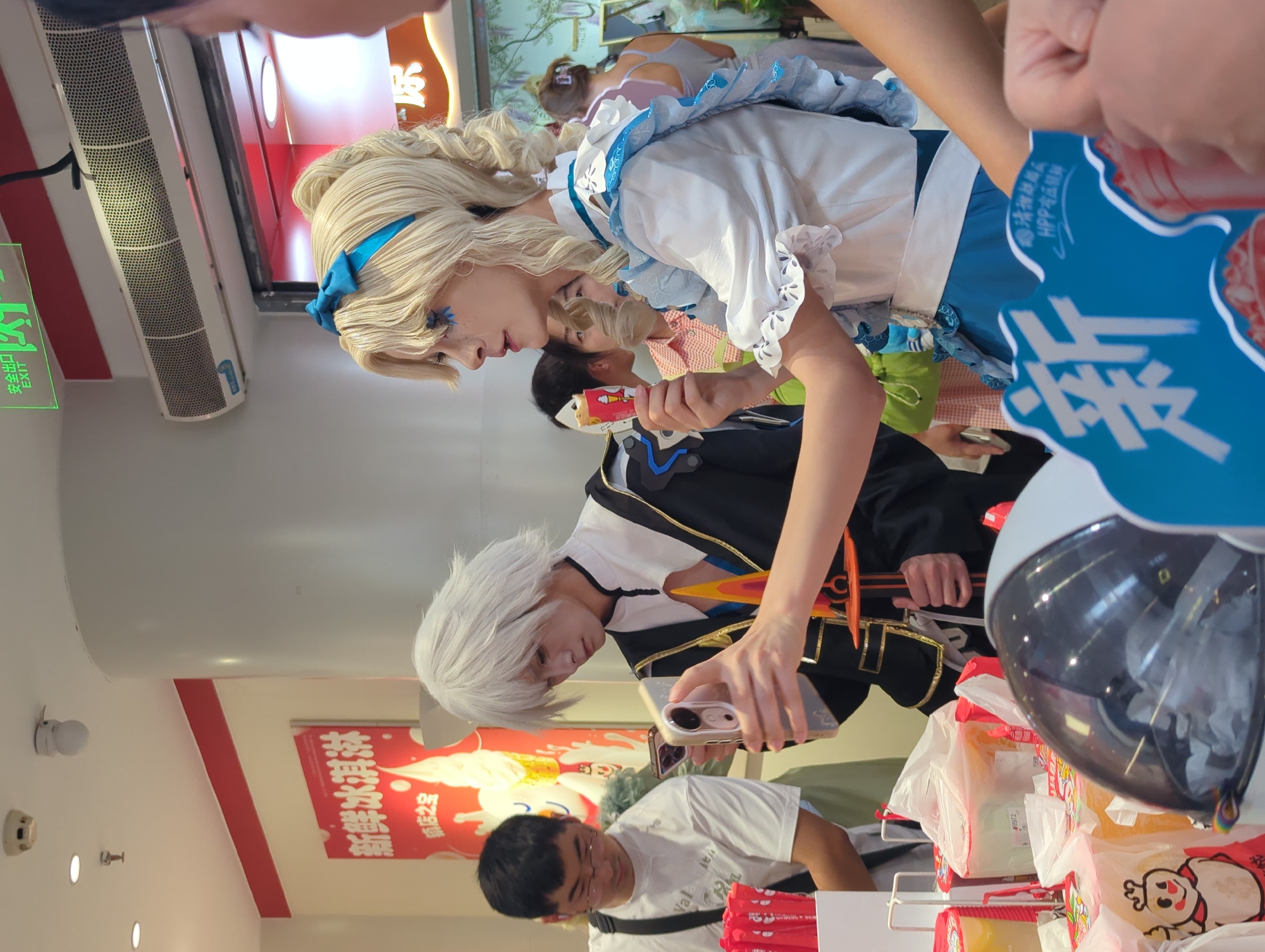In Qingyang District, China, a group of people are gathered in what appears to be a store, possibly a bubble tea shop or similar establishment, indicated by the "MIXUE ICE CREAM & TEA" branding visible on red packaging. The foreground is dominated by a large, spherical, transparent display case, possibly for ice cream or beverages. To the left, a man wearing glasses and a white t-shirt is visible. In the center, a person in an elaborate blonde wig and a blue and white costume, likely a cosplayer, is holding a phone and an ice cream cone. This individual has blue makeup around their eyes and is looking to the left. Another person in a white wig and a black and gold outfit, also possibly a cosplayer, stands to the left of the blonde individual, holding a phone and a prop sword. Behind them, other individuals are visible, including a woman in a pink and white checkered shirt and another in a gray tank top. The background features store signage, including a red sign with Chinese characters that reads "新鲜冰淇淋" (Fresh Ice Cream) and a green "EXIT" sign. The environment appears to be an indoor commercial space.