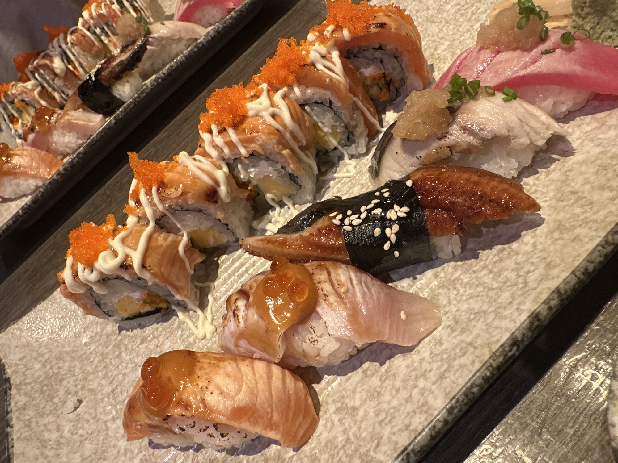 A plate of sushi is depicted, showcasing various types, including salmon rolls with orange roe and mayonnaise drizzle, eel nigiri with sesame seeds, and other nigiri with different toppings. Another plate of similar sushi varieties is partially visible in the background. The image does not contain information about the location or if it's in บ้านบางรักษ์, Thailand.