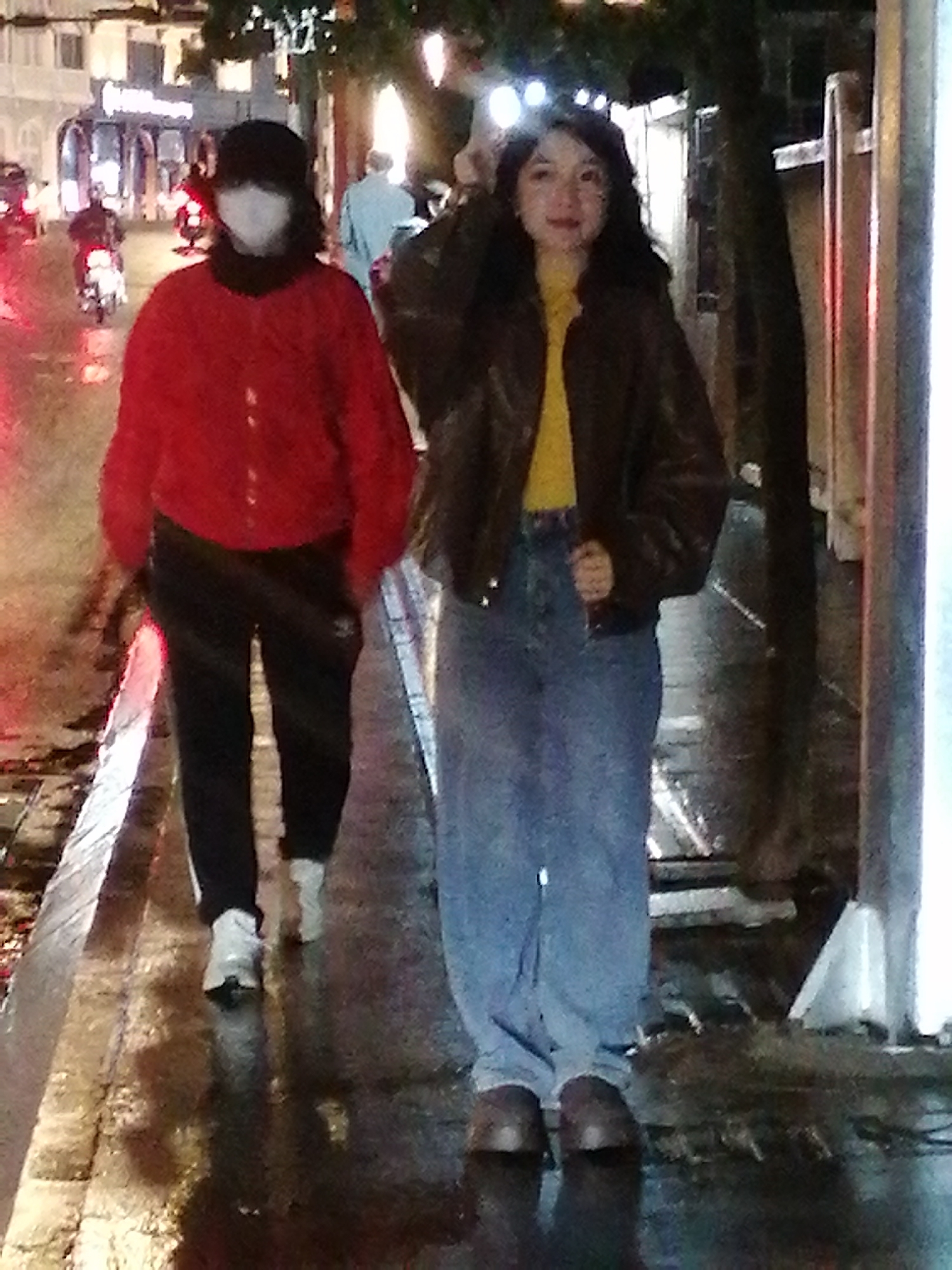 The image depicts two individuals walking on a wet, paved sidewalk at night in Da Lat, Vietnam. The person in the foreground on the left wears a red jacket, black pants, and white sneakers. They have a black hat and a white face mask covering their face. The person in the foreground on the right wears a brown leather jacket over a yellow shirt, blue jeans, and dark shoes. They have dark hair and appear to be looking forward. The background shows a street with vehicles, including motorcycles, and illuminated buildings. The overall atmosphere suggests a busy urban environment at night, possibly during or after rain, given the wet surfaces.