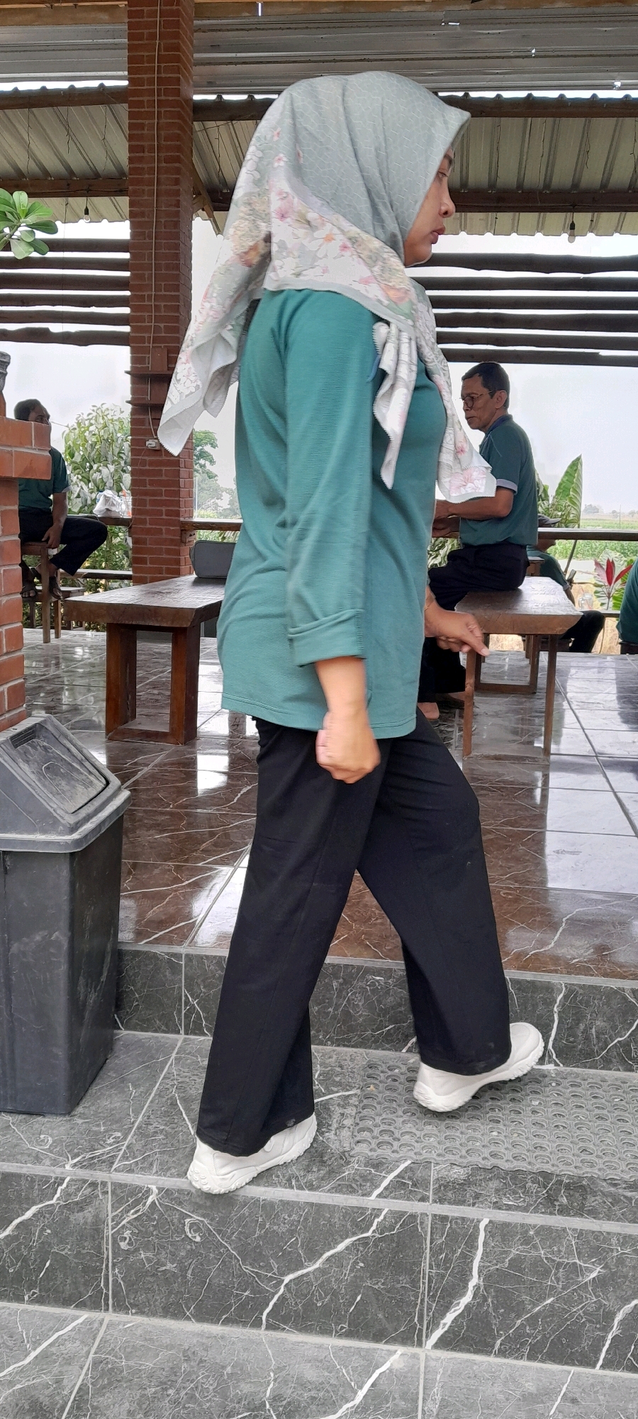 In Sidohasri, Indonesia, a woman wearing a teal long-sleeved shirt, black pants, white sneakers, and a light blue hijab with floral patterns is walking on a stone tiled staircase. Her right hand is clenched in front of her. Behind her, a black trash can is visible on the left side of the frame. In the background, two men are seated at wooden tables under a covered outdoor seating area with a corrugated metal roof supported by brick pillars. One man, wearing glasses and a green shirt, is seated on a wooden bench to the right. The other man, wearing a green shirt and black pants, is seated on a stool on the left. The environment appears to be a semi-outdoor dining or resting area.