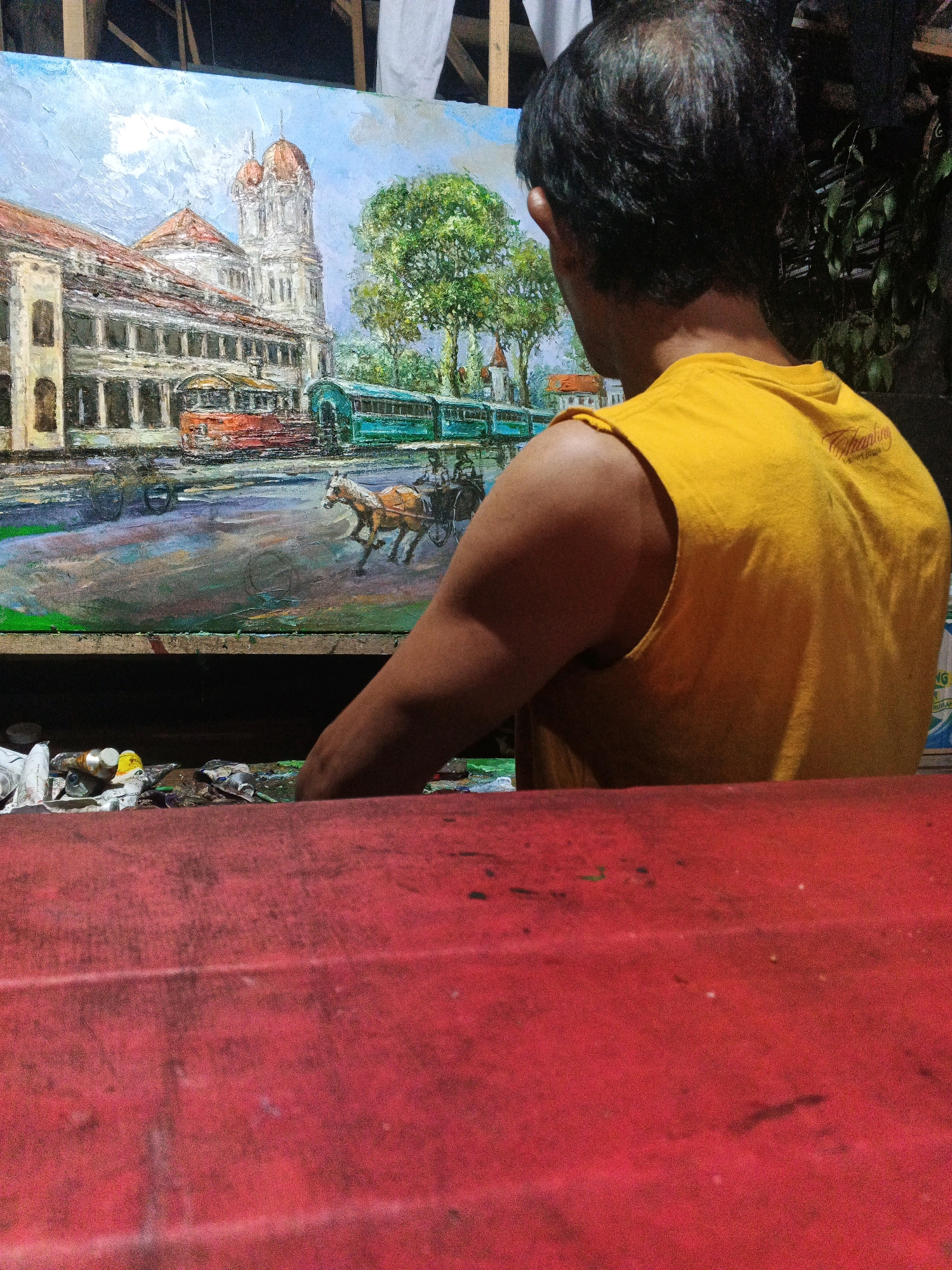A man in a yellow tank top stands with his back to the camera, looking at an oil painting. The painting depicts a street scene in Ungaran, Indonesia, with a colonial-era building and two trams. A horse-drawn carriage is in the foreground. The man's right arm is raised and his hand is reaching towards the painting. In front of the painting, on a red surface, are tubes of paint and brushes. The background is dark and out of focus, with some greenery visible.