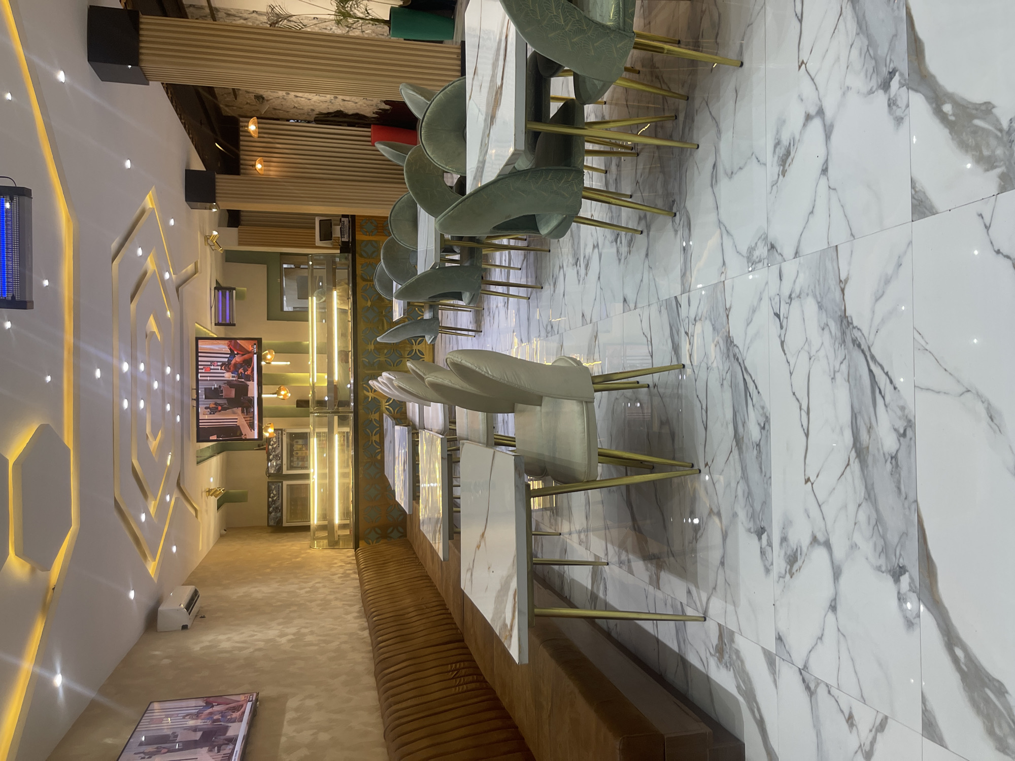 The image shows the interior of a modern restaurant in Jos, Nigeria. The restaurant features a long, tufted brown banquette along the left wall, facing a row of white marble-top tables with gold-colored legs. Several light-colored chairs with gold legs are positioned around the tables. The floor is covered in large white marble tiles with grey veining.
In the background, there is a display counter with illuminated shelves, possibly for food or desserts. Two large flat-screen televisions are mounted on the wall. One is on the left, displaying an image of people, and another is further back on the right. The ceiling has a modern design with recessed lighting and yellow accents. Wooden pillars with vertical slats are visible on the right side of the image. The overall ambiance is clean, contemporary, and well-lit. There are no people visible in the image.