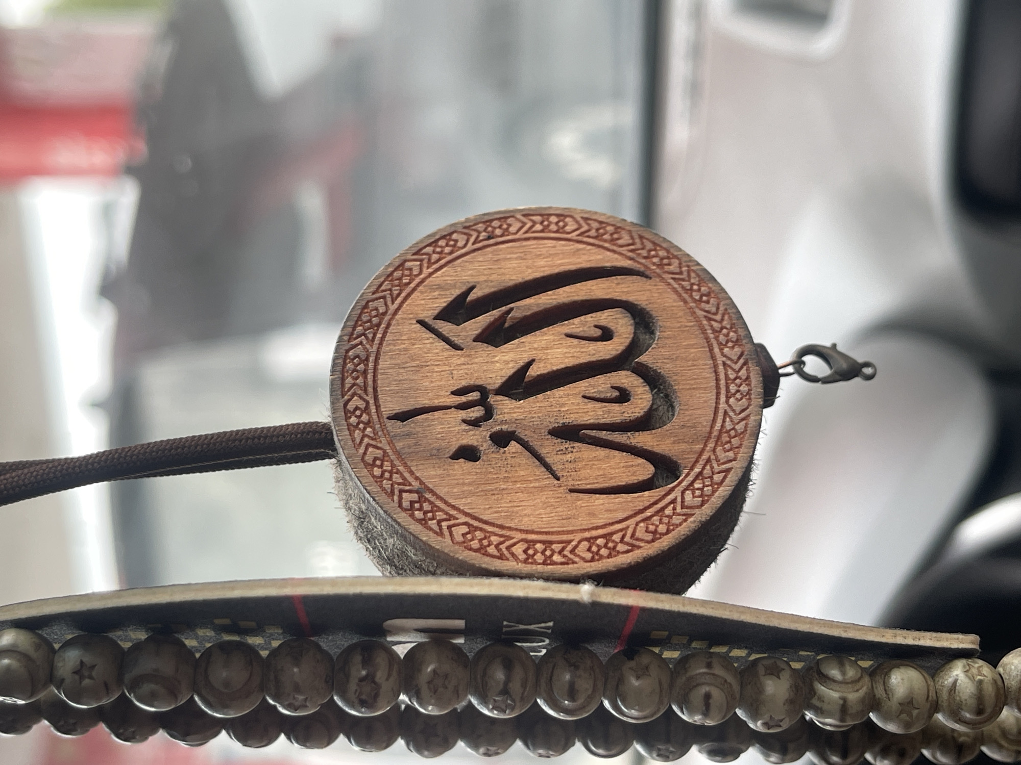 A close-up shot focuses on a circular wooden pendant with Arabic calligraphy carved into it, set against a blurred background. The pendant has a decorative border with a geometric pattern. It is attached to a dark brown cord and a small metal clasp. Below the pendant, a row of light-colored beads, some carved with star shapes, are visible, resting on a dark, flat surface. The scene appears to be indoors, possibly in a vehicle or near a window, given the reflections and blurred light in the background. The overall mood is calm and contemplative. The location is Istra, Russia.