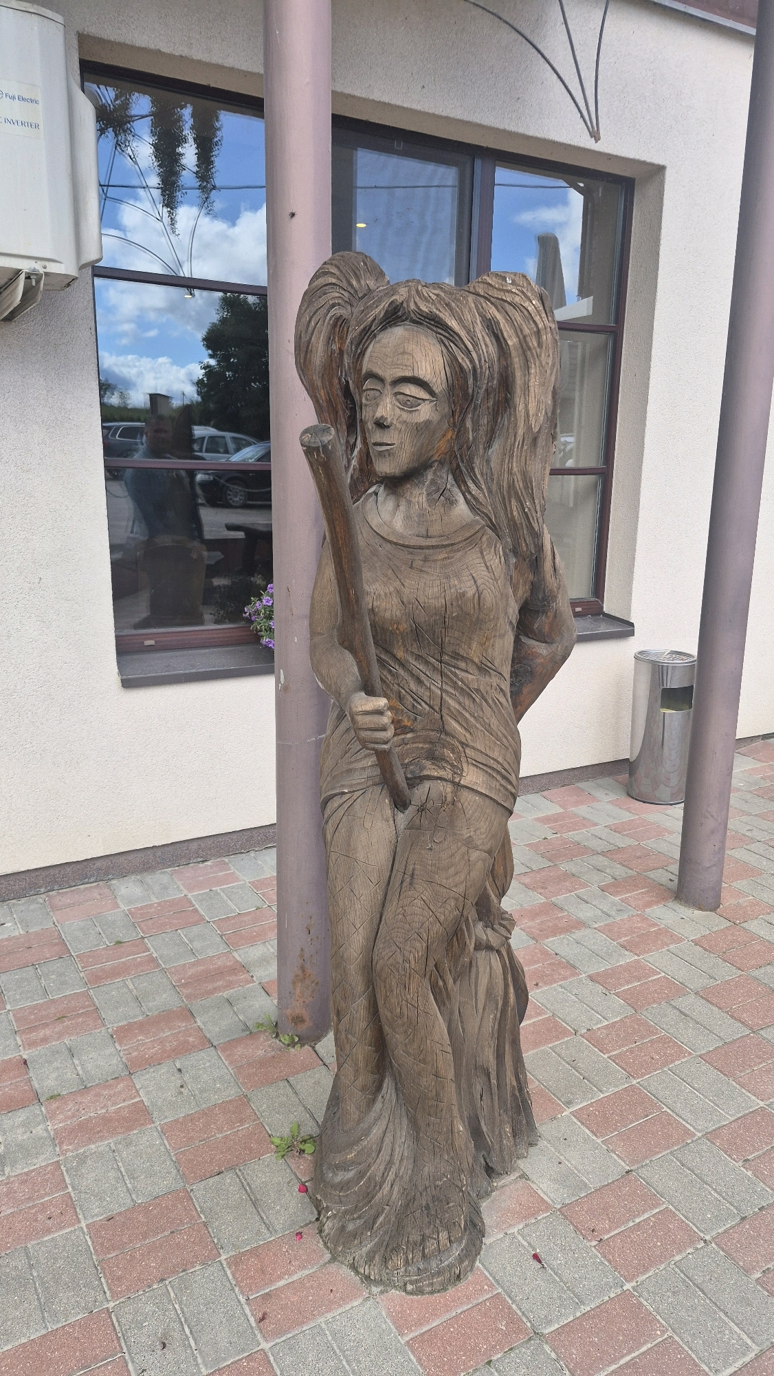The image shows a carved wooden sculpture of a woman with large pigtails, holding a staff. She is standing in front of a building with large windows that reflect the sky and trees. The sculpture is placed on a patterned brick walkway. To the right of the sculpture is a metal trash can. To the left, an outdoor air conditioning unit is visible. The overall environment appears to be an urban or semi-urban setting in the city of Smėlynė, Lithuania. The wooden sculpture is aged, with visible cracks and texture. The lighting suggests it is daytime, with a mix of direct and reflected light.