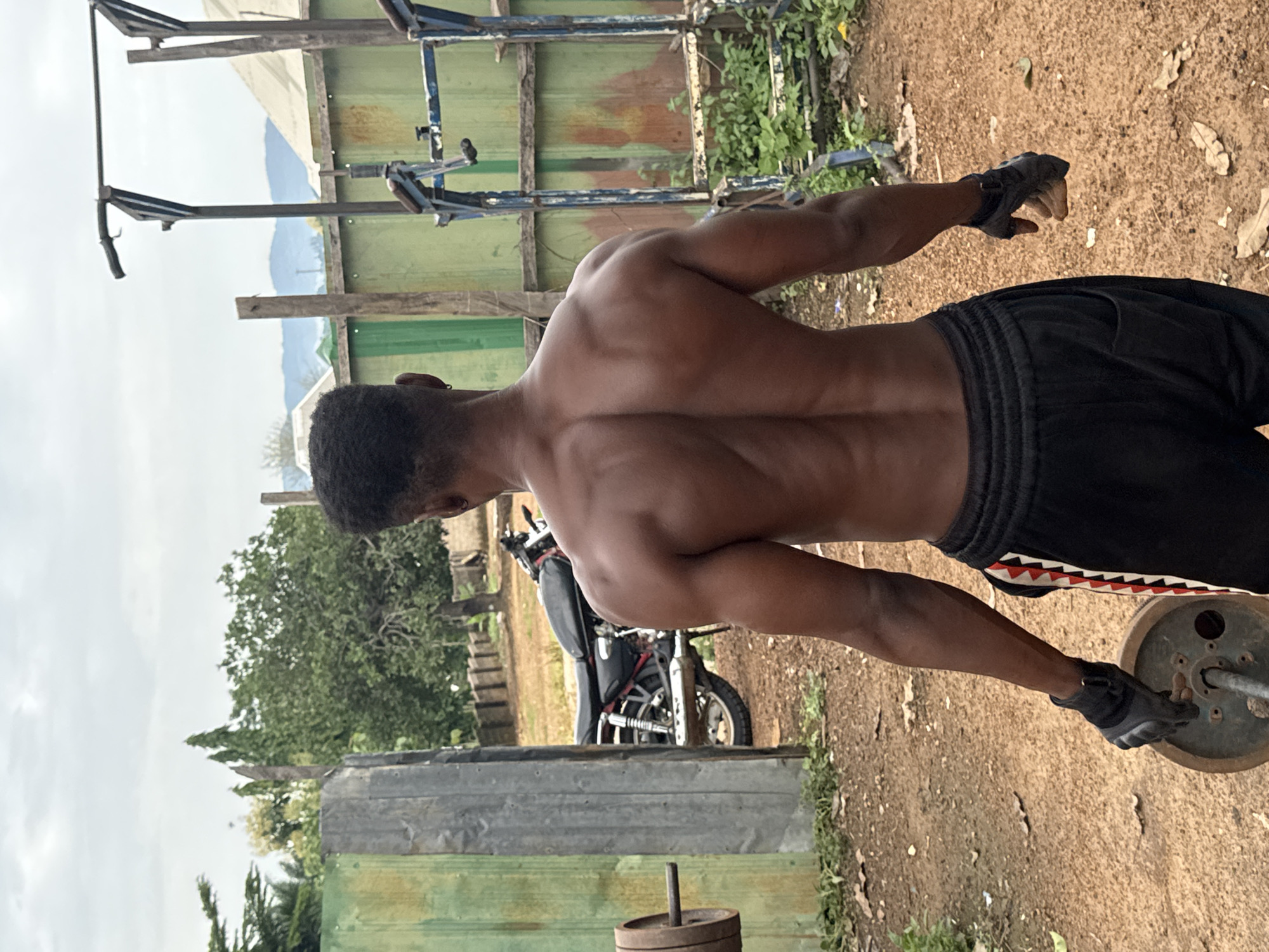 A shirtless Black male is standing outdoors, facing away from the camera, in what appears to be an outdoor gym in Akwanga, Nigeria. He has dark skin, short dark hair, and a muscular physique, with his back muscles prominently defined. He is wearing black athletic shorts with a red and white striped pattern on the side and black weightlifting gloves on both hands. In his left hand, he is holding a barbell weight.
The background consists of a dirt ground, some greenery, and a green corrugated metal fence. Behind the fence, a motorcycle is parked. In the distance, there is a rudimentary weightlifting apparatus, likely a pull-up bar or squat rack, and hills can be seen under a cloudy sky. The overall scene suggests a casual, perhaps makeshift, outdoor workout environment.