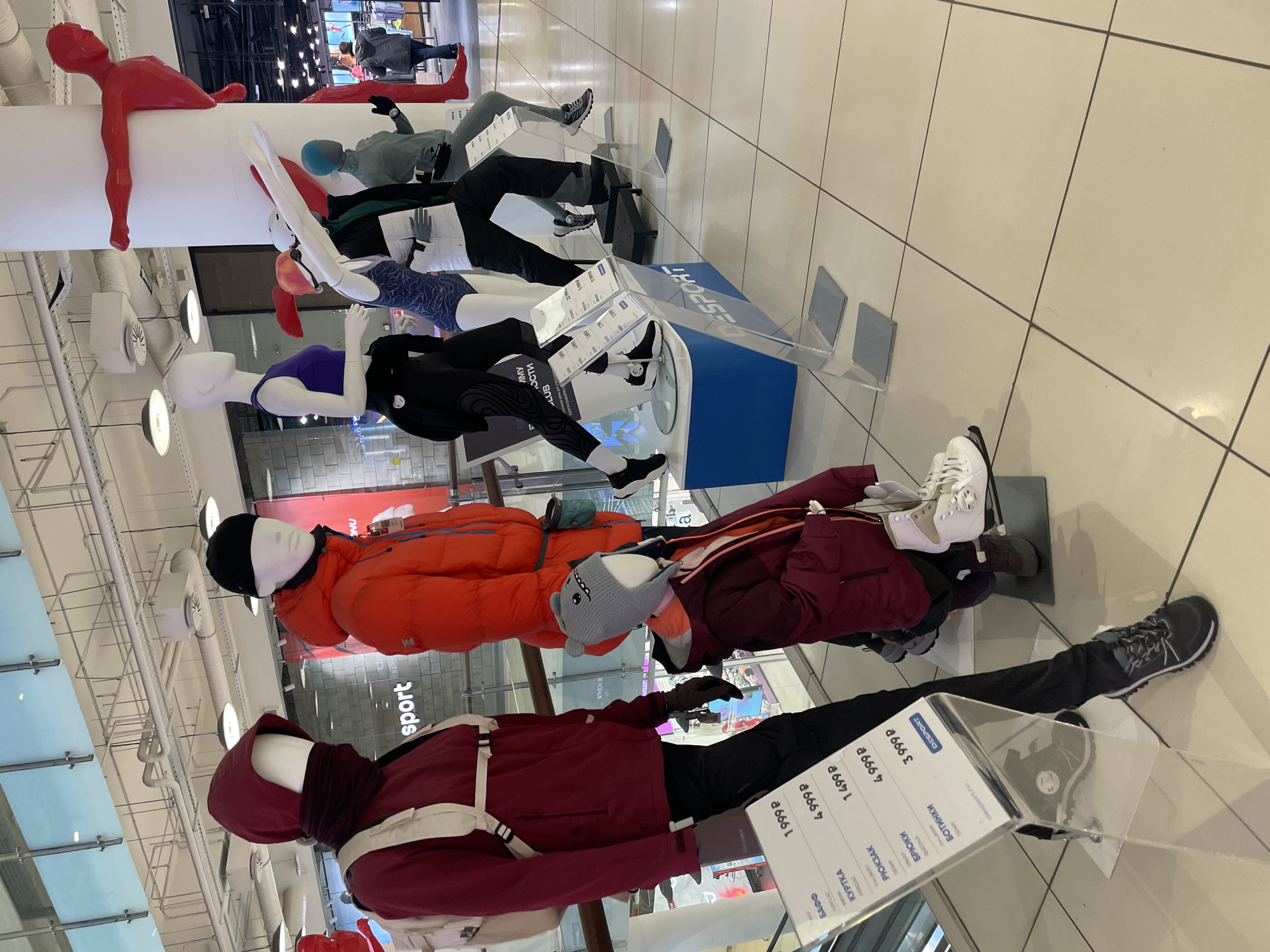 The image depicts a retail store display, likely located in Khimki, Russia, featuring mannequins dressed in athletic wear. The mannequins are arranged in a row and showcase a variety of outdoor clothing and accessories, including jackets, pants, and shoes. A column with figures on it hangs from the ceiling. Clear plastic display stands with price tags are placed in front of some of the mannequins. The image is taken at an angle, providing a perspective from the floor level. The setting appears to be a modern retail environment with tiled flooring and bright lighting. The price tags contain Cyrillic text, confirming the location as likely being in Russia.