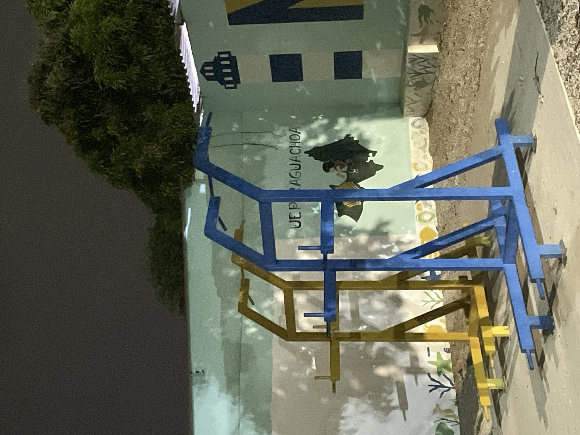 The image shows a blue and yellow metal playground structure situated in front of a painted wall. The wall has a light green background and features various graphic elements, including a lighthouse, blue and black squares, a map of Paraguay with the words "UE P RAGUACHOA" written above it. At the base of the wall, there are images of plants and marine life. To the left of the wall, there is dark foliage, likely a bush or tree. The ground to the right of the playground is concrete. The scene appears to be located in Paraguay, as indicated by the map painted on the wall.
