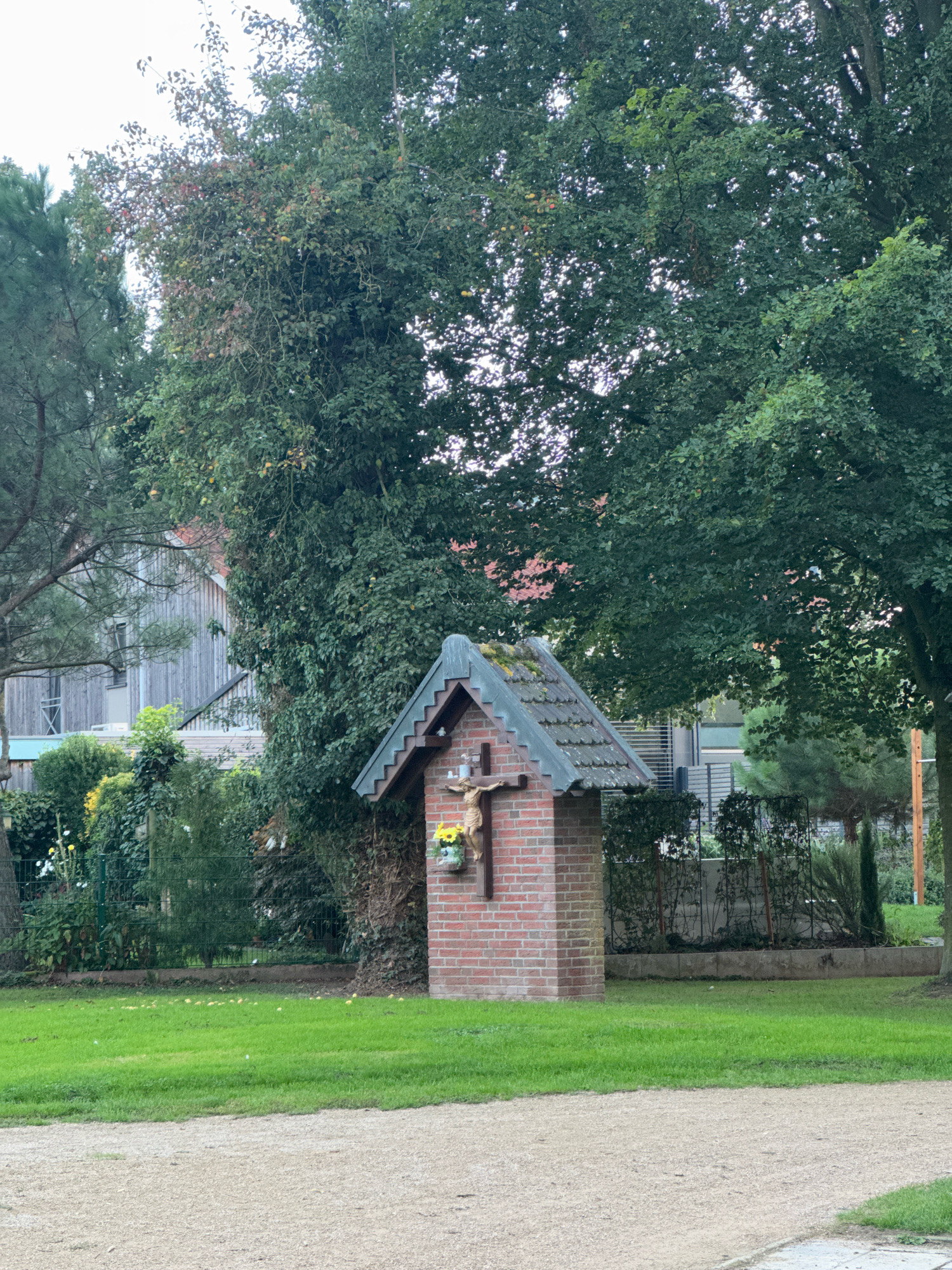 In the city of Cleves, Germany, a small brick shrine stands in a grassy area. The shrine is topped with a grey, tiled roof and features a crucifix with a figure of Jesus attached to its front. Yellow flowers are placed at the base of the crucifix. Behind the shrine, a large, leafy tree dominates the scene, with its branches extending upwards. To the left of the shrine, a wooden building with a grey facade and a pitched roof is visible. The ground in front of the shrine is covered with gravel, and a green lawn stretches out behind it. The environment suggests an outdoor setting, possibly a park or a garden, with a mix of natural and man-made elements.