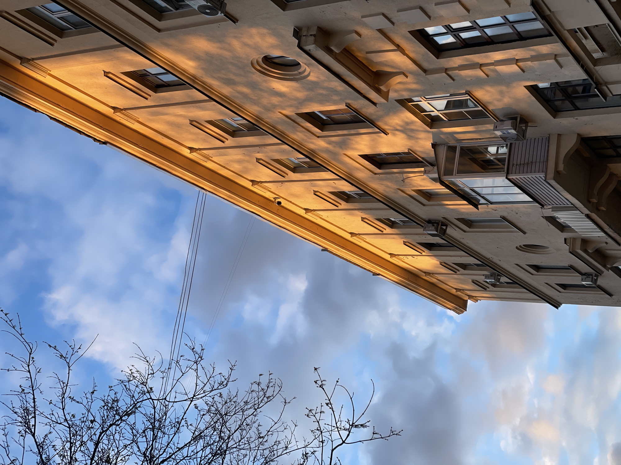 The image presents an upward perspective of a multi-story building facade under a dynamic sky, with bare tree branches in the foreground. The scene is set in Khimki, Russia.
The building, likely of classical or neoclassical architectural design, features ornate details such as cornices, rectangular window frames, and at least one prominent circular decorative element on its beige-colored exterior. A warm, golden light, suggesting either sunrise or sunset, illuminates the upper portions of the facade, creating distinct shadows from windows and architectural protrusions across its surface.
The sky is a blend of clear blue patches and soft, scattered white and light grey clouds. Overhead, thin utility wires cut across the upper left quadrant of the frame. In the lower left, a cluster of leafless tree branches, indicative of winter or early spring, is silhouetted against the sky. Some windows on the building reflect the sky and clouds. A small, glass-enclosed balcony or extension protrudes from one section of the facade, and external units, possibly air conditioning units, are visible.
There are no people or specific activities depicted. No text is visible. The notable details include the striking interplay of golden light and shadow on the architectural elements, the starkness of the bare tree branches, and the contrasting textures of the urban structure against the natural sky.