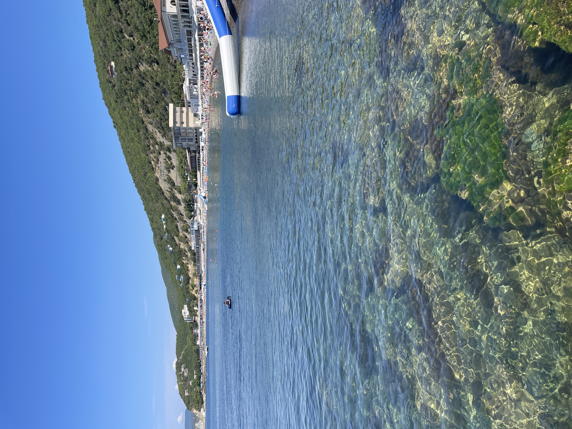 The image depicts a sunny day at a beach in Shirokaya Balka, Russia. In the foreground, clear turquoise water reveals the rocky seabed covered with green algae. Further out, the sea is calmer, with several people visible. One person is lying on an inflatable raft. The background features a densely vegetated hillside with buildings scattered along its slope and at its base. A long stretch of beach with numerous umbrellas and sunbathers lines the shore, backed by a row of white buildings, likely hotels or resorts. An inflatable water slide is partially visible extending into the water from the right side of the image. The sky is a clear, vibrant blue.