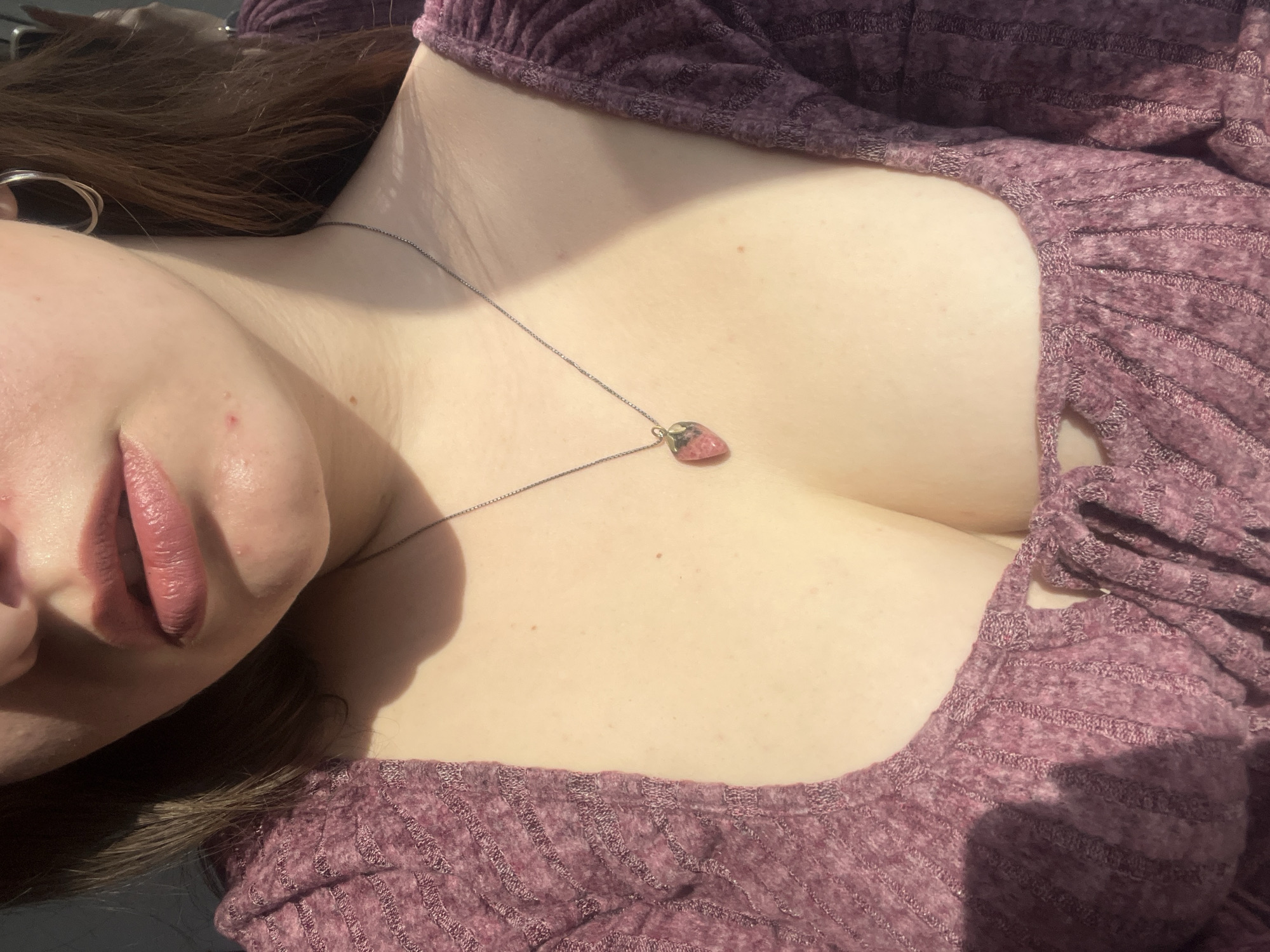 An eye-level, close-up shot shows a woman's face and upper chest angled downward. Her full lips are visible, colored with a mauve shade of lipstick. She wears a burgundy-colored, ribbed top with a low, gathered neckline that reveals cleavage. A thin, silver chain necklace hangs around her neck, suspending a small, pink stone pendant. The woman's hair is dark brown. A silver hoop earring is partially visible in the upper left corner. The background is out of focus. The location cannot be determined from the image.