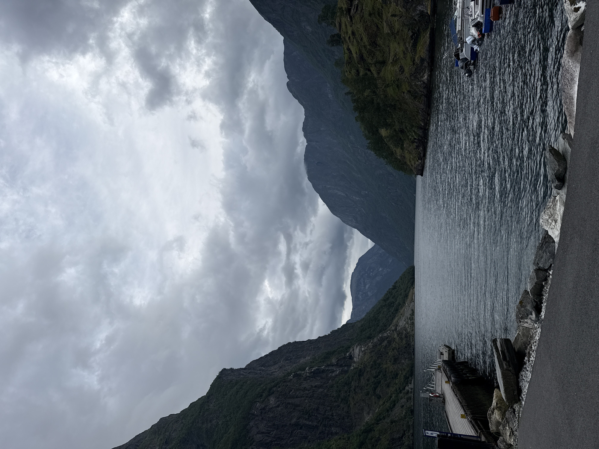 This is a wide, landscape photograph of the Gudvangen fjord in Gudvangen, Norway. The image is dominated by dark, imposing mountains that rise steeply on either side of a tranquil, dark body of water. The sky is overcast with heavy, grey clouds, creating a somber and dramatic atmosphere. In the foreground, a wooden pier juts out into the water, with a few small boats moored alongside it on the right. A red life preserver hangs on the pier. The shoreline on the left is a mix of rocks and a paved path. The overall impression is one of natural grandeur and a quiet, somewhat isolated, coastal setting.