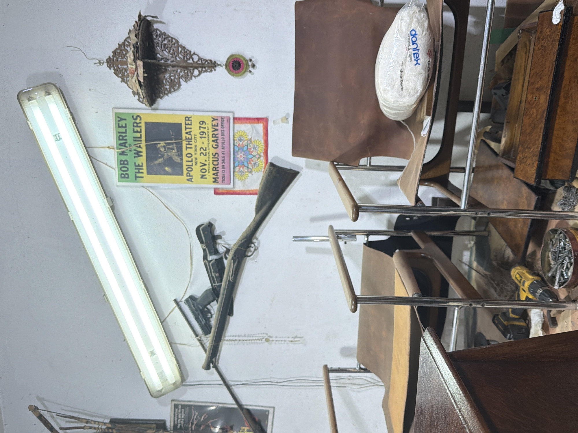 A fluorescent light fixture illuminates a crowded interior scene with a light-colored wall. Affixed to the wall are various items including a metal decorative element, a colorful circular decoration, a Bob Marley and the Wailers concert poster from the Apollo Theater in New York City dated November 22, 1979, and a patterned textile. Leaning against the wall is a long gun or rifle, partially obscuring a black and white image and other objects. In the foreground, several brown leather chairs with metal frames are visible, with a bag labeled "dontex absorbent" resting on one. Tools, including a yellow power drill and a container of screws, are situated on a shelf or surface near the chairs.