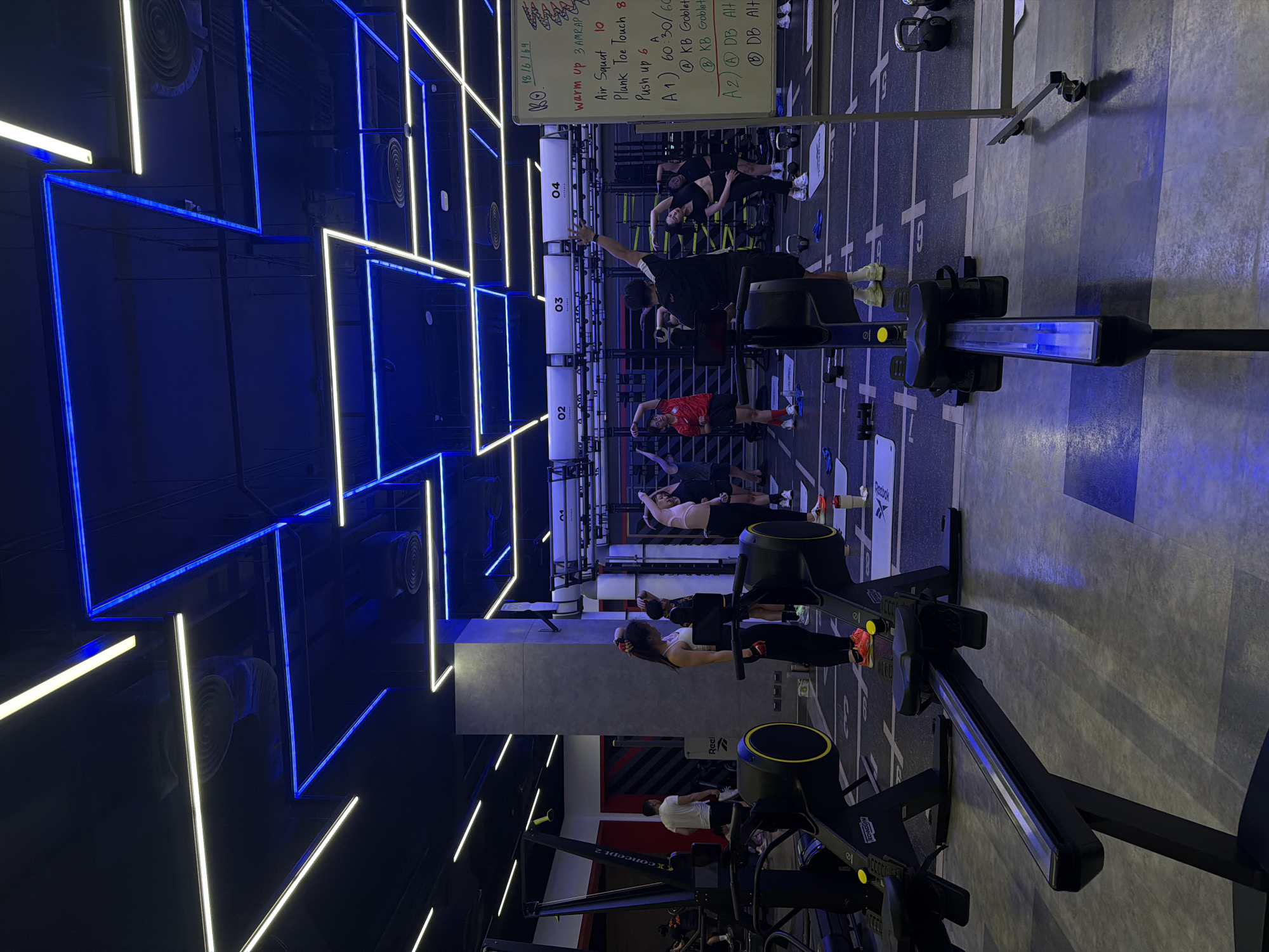 The image depicts an indoor fitness studio with a group of people participating in a workout session. The environment is dimly lit, with prominent blue and white LED strip lighting illuminating the ceiling and surrounding areas. The floor is a patterned grey tile, marked with white lines and squares, suggesting designated workout stations.
In the foreground, two rowing machines are visible, with one partially obscuring the other. These machines are black with yellow accents. The further rowing machine has a blue light illuminated on its central console.
In the mid-ground, several individuals are engaged in stretching or exercise routines. One woman in the center foreground, wearing a white sports bra and black leggings, stands facing a tablet device. To her right, several individuals are performing side stretches, with one woman in a pink sports bra and black leggings and another in a black sports bra and shorts. Behind them, a man wearing a black t-shirt and shorts appears to be instructing the group, gesturing with his right arm. A person in a red t-shirt and black shorts is also visible.
In the background, a large whiteboard displays workout instructions, including sections for "WARM UP", "Air Squat", "Plank Toe Touch", and "Push up". Numerical sets and repetitions are listed alongside exercises labeled "A1", "A2", "B", and "DB". There are additional people further in the background, and various gym equipment, including dumbbells and kettlebells, are scattered on the floor. The overall impression is that of a structured fitness class.
The location is identified as a fitness studio in Nong Pa Khrang, Thailand.