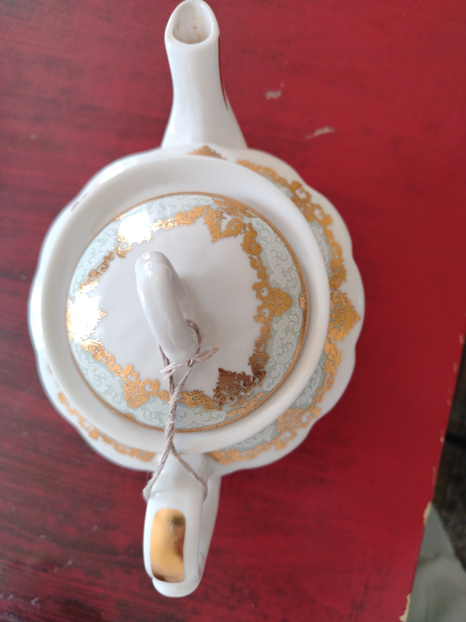 An overhead shot shows a white porcelain teapot with gold trim and delicate light blue patterns. The teapot is placed on a dark red wooden surface. A thin twine is tied around the handle of the lid.
