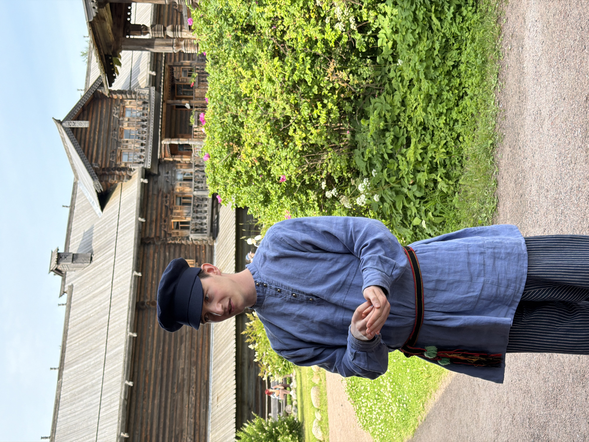 A young man stands in front of a wooden building, wearing a blue, long-sleeved shirt and a dark blue cap. He has his hands clasped together in front of him. A colorful sash is tied around his waist, with red, black, and green threads. He is wearing striped pants. Behind him, a large, ornate wooden structure with multiple levels and balconies is visible. Lush green bushes and some small white flowers are in the foreground, partially obscuring the lower part of the wooden building. The ground is a gravel path. The sky is a pale blue with a few wispy clouds. The overall setting appears to be an outdoor historical or cultural site. The location is the Museum of Wooden Architecture in Imeni Sverdlova, Russia.