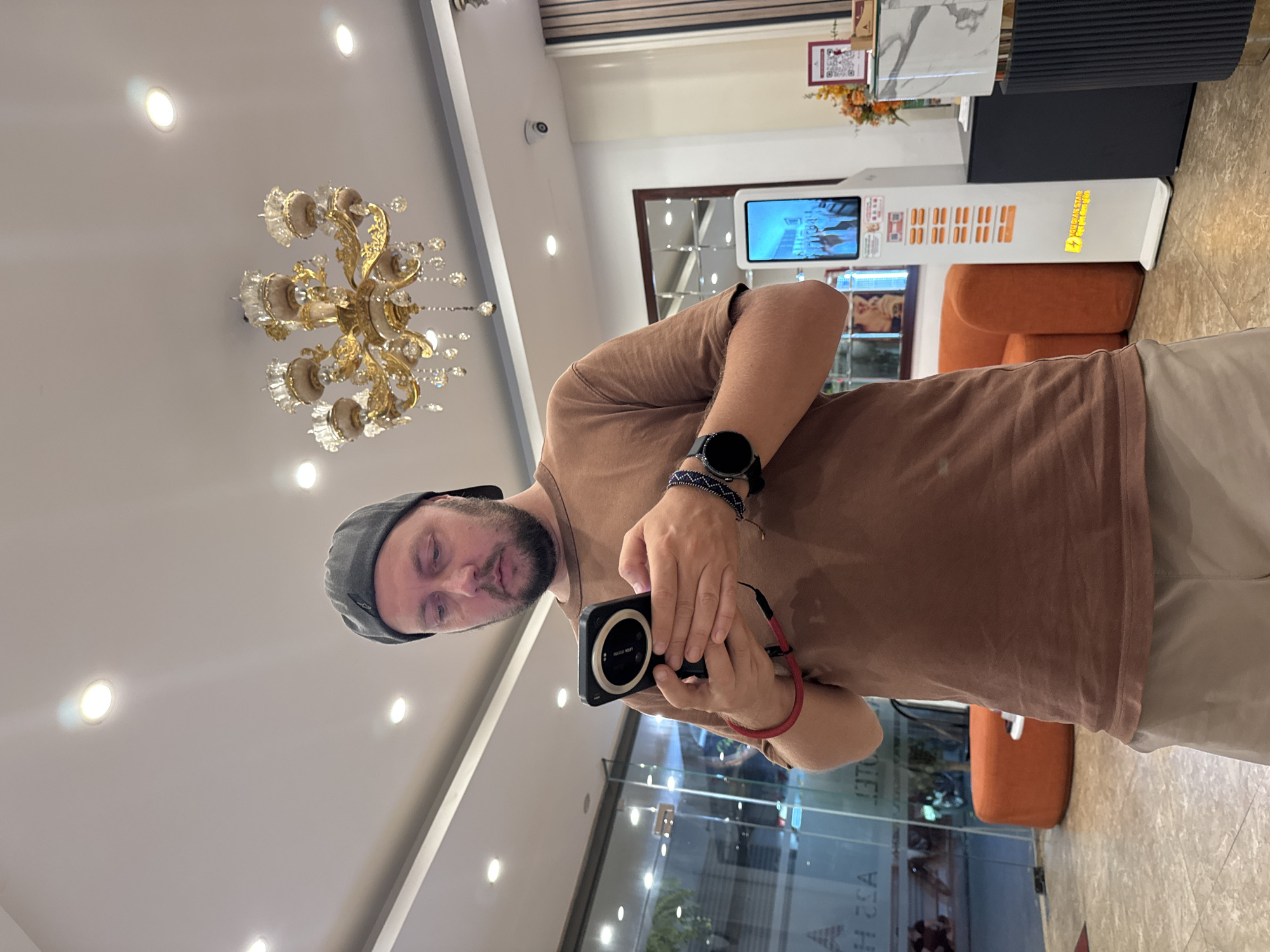 This is a selfie taken by a man wearing a grey baseball cap, a brown t-shirt, and light-colored shorts. He is holding a smartphone in his right hand, which has a red strap attached. He is also wearing a smartwatch on his left wrist. The man is standing in what appears to be an indoor lobby area, possibly a hotel or a commercial building.
In the background, there is a large, ornate gold chandelier hanging from the ceiling, illuminated by several recessed ceiling lights. To the right, a digital display screen is visible, showing images or text. Further back, an orange armchair is placed near a counter. The floor is tiled. The overall ambiance suggests a well-lit and modern indoor space. There is some text visible on the digital display, but it is not clearly legible. The image is oriented vertically, as if the photo was taken with the phone held in a portrait orientation and then rotated. The lighting suggests it could be daytime, though no direct sunlight is visible. The man's facial expression appears neutral. The location context provided is Hanoi, Vietnam.
