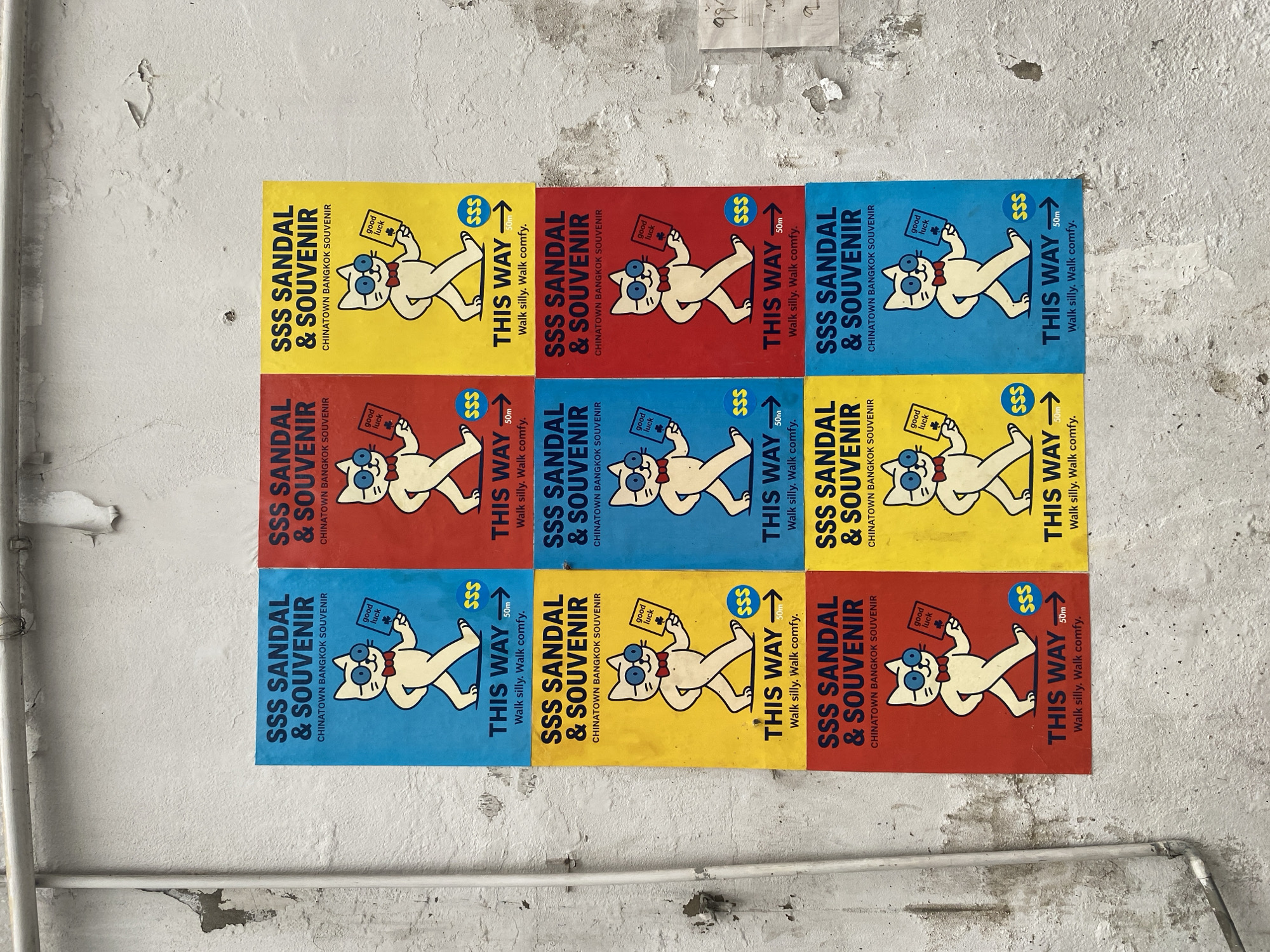 The image shows a close-up of a section of a wall in Chinatown, Bangkok, Thailand. Nine advertising posters are arranged in a 3x3 grid. Each poster is rectangular and features a cartoon cat wearing glasses and a bow tie, holding a "good luck" card. The text on each poster reads "SSS Sandal & Souvenir, Chinatown Bangkok Souvenir, This Way 50m Walk silly. Walk comfy." The posters vary in color. The background is a weathered wall, showing signs of age and wear. A white pipe runs vertically along the left side.