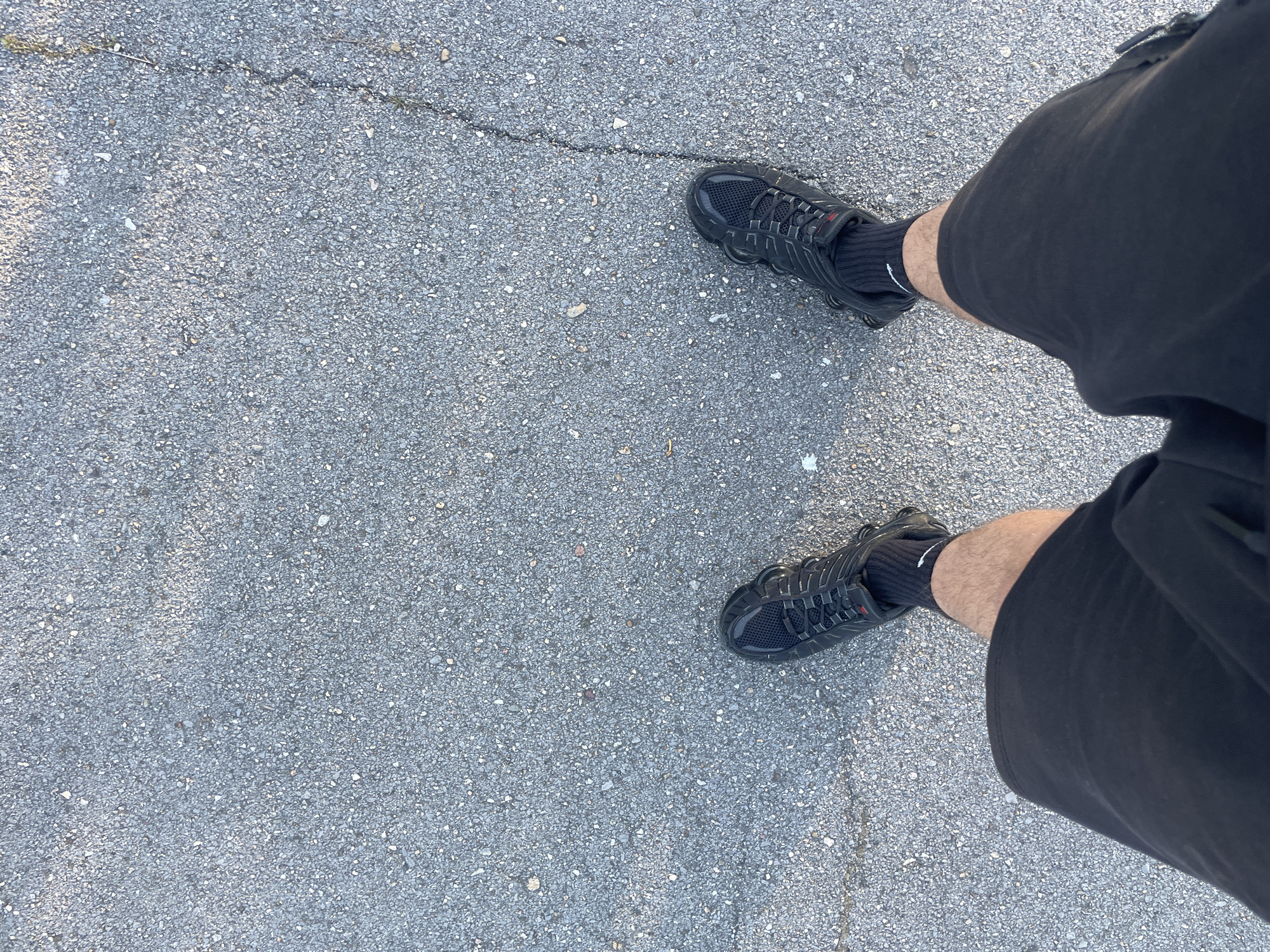 The image, taken from a top-down perspective, features the lower half of a person's body standing on an asphalt surface. The subject is wearing black shorts and black athletic shoes. The shorts cover the upper thighs, revealing bare skin on the lower thighs and calves. The shoes are black sneakers with a distinctive design, including visible segmented soles and a textured mesh upper. Black socks are worn with the sneakers; the left sock has a small white logo, likely a Nike swoosh, visible on the outer ankle.
The ground is a grey, textured asphalt or similar paved surface, consistent with an outdoor urban environment like a street or sidewalk. There are minor cracks in the pavement, with a more prominent, dark crack running diagonally across the upper central portion of the image. The lighting suggests it is daytime, with bright sunlight creating subtle shadows. The overall setting is simple, focusing on the person's stance on the ground. The location is Sofia, Bulgaria.