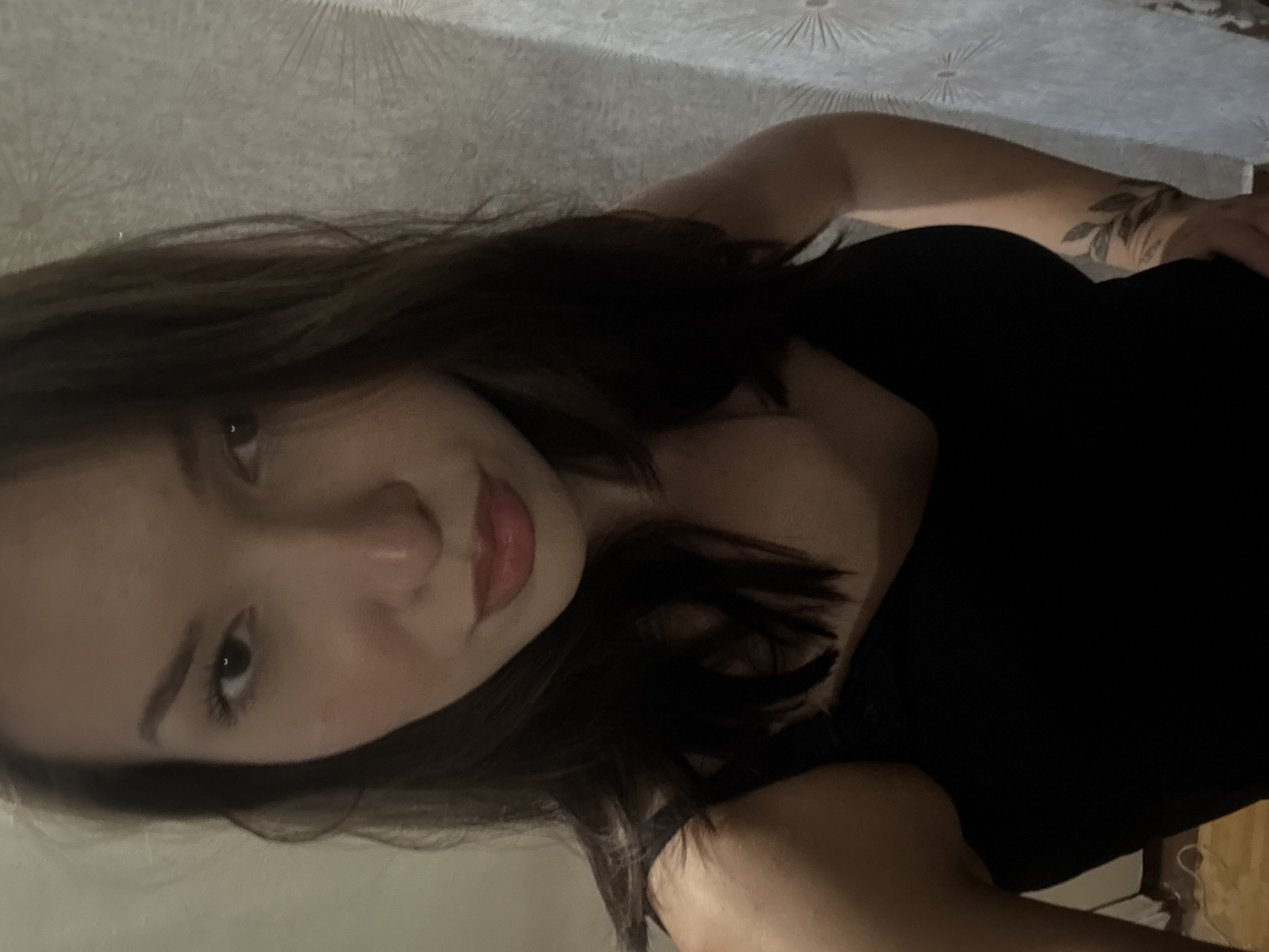 A young woman with dark hair, wearing a black tank top, poses for a selfie in an indoor setting. The background consists of a patterned wallpaper and a partially visible white curtain. A subtle leaf tattoo is visible on her arm. The lighting is dim, casting shadows on her face and body. The woman is positioned in the foreground, with her head slightly tilted and her lips slightly parted. Her gaze is directed towards the camera. The location is specified as the city of Inskoy, Russia, though there are no specific visual cues in the image to confirm this.