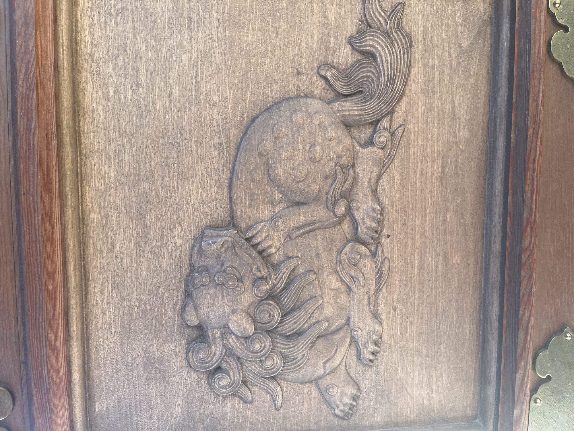 The image shows a close-up of a wooden carving of a mythical creature, possibly a Komainu or Foo dog, on a wooden surface. The carving is detailed, with swirling mane and stylized paws and tail. The wood appears to be aged and has a visible grain. The carving is set within a frame of darker wood at the top and bottom, with what appear to be brass decorative elements at the bottom corners. The lighting suggests the image was taken in daylight, casting subtle shadows that highlight the carving's relief. There is no indication of the city of Krasnodar, Russia, or any specific location.