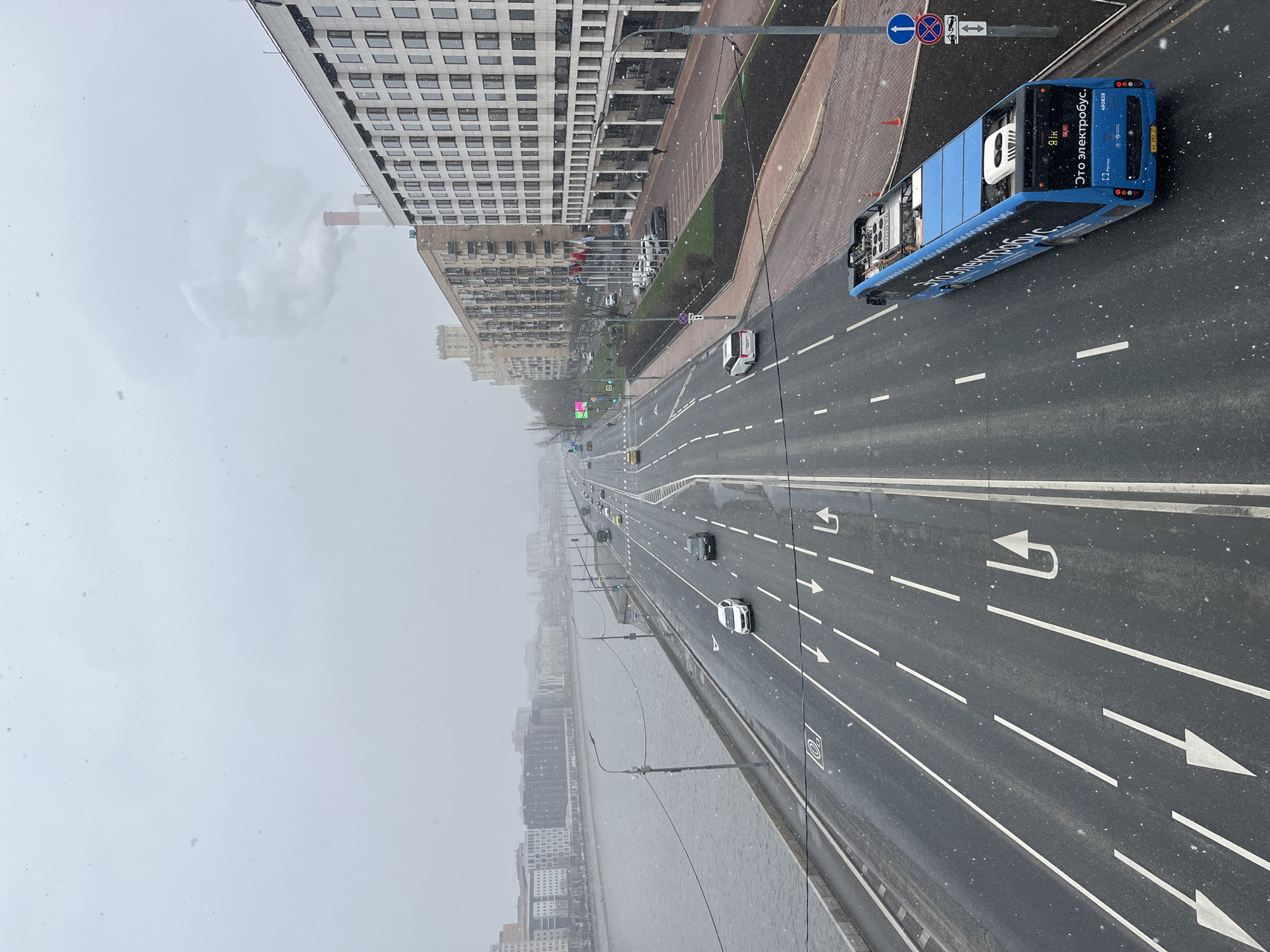 The image depicts an elevated view of a major roadway in Moscow, Russia, during a period of snowfall. The road has multiple lanes delineated by white lines and arrows indicating direction. A blue electric bus ("Это электробус") is visible on the right-hand side of the road. There are several cars driving along the road. Along the side of the road is a row of buildings, including a high-rise structure with a repetitive window pattern. A river flows alongside the road on the left side of the image. Street signage is present at the intersection on the right side of the image. The overall weather condition is snowy and overcast.