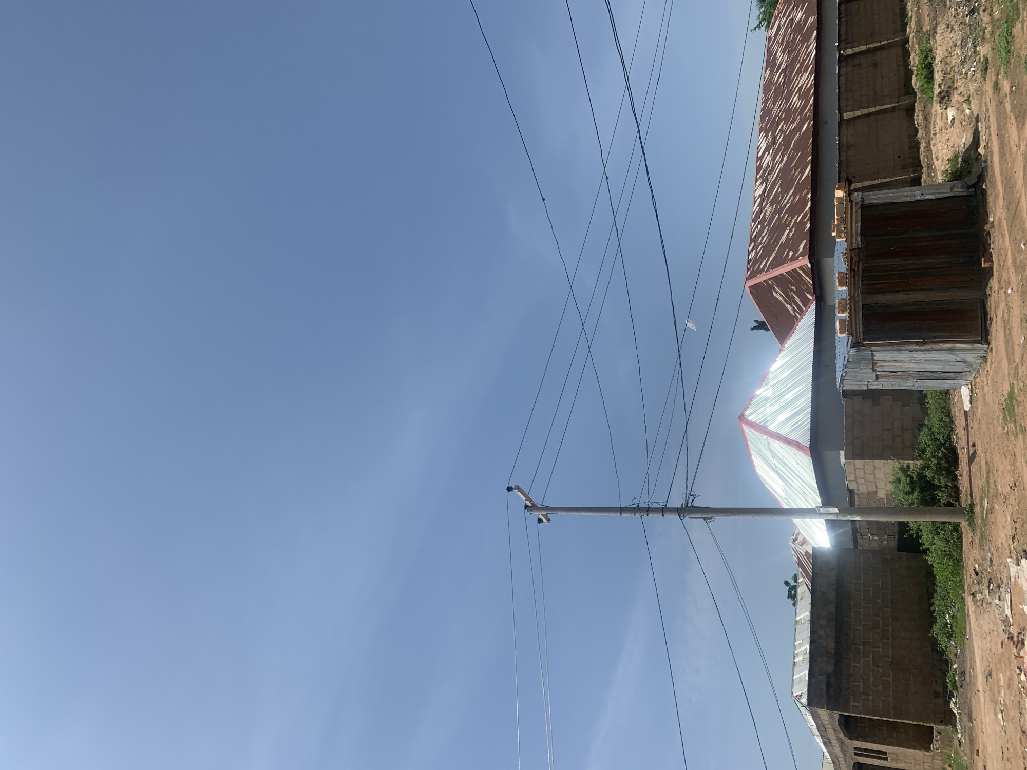 In the city of Jauro Yinu, Nigeria, this image captures a street-level view under a clear, bright blue sky. The foreground consists of a dirt road littered with debris, including pieces of paper and what appears to be wood. To the left, a building constructed with mud bricks is partially visible, featuring an open doorway and visible windows. Adjacent to this is another structure with a metallic, red-trimmed roof. Further to the right, a small, weathered corrugated metal shed stands, topped with loose bricks. Behind the shed, a long, low wall made of mud bricks extends, and beyond that, a larger building with a rusted, red corrugated metal roof is prominent. A utility pole with multiple overhead wires runs through the mid-ground, cutting across the scene. The overall environment suggests a rural or semi-urban setting in Nigeria, characterized by simple construction and open, dusty terrain.