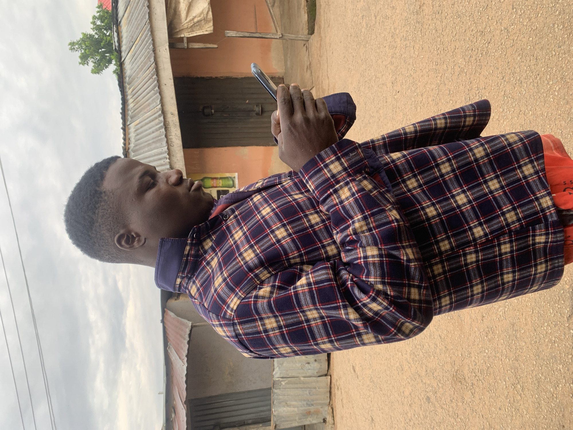 A male individual, likely an adult, is depicted outdoors in a candid, waist-up shot. He possesses a dark complexion and short, dark hair. He is wearing a long-sleeved plaid shirt, primarily dark blue or purple, with patterns of tan, red, and light blue lines. A dark blue collar is visible. A red garment is worn underneath, partially visible at the bottom of the frame. The man is holding a silver-backed smartphone horizontally with both hands, looking intently downwards at the screen.
The setting appears to be an unpaved area in Jos, Nigeria, characterized by light brown/tan ground with small pebbles. In the background, an overcast, light grey sky is visible. Behind the man, an orange-colored single-story building with a dark wooden door and a corrugated metal roof is prominent. A green bottle advertisement or poster is affixed to the wall to the left of the door. Faint, handwritten text is visible on the corrugated roof above the door, including "OPO 360270" and "Tk 0808...". To the far left, another structure with corrugated metal siding and roof is partially visible. A portion of a green tree is seen above the corrugated roof on the right side of the frame.