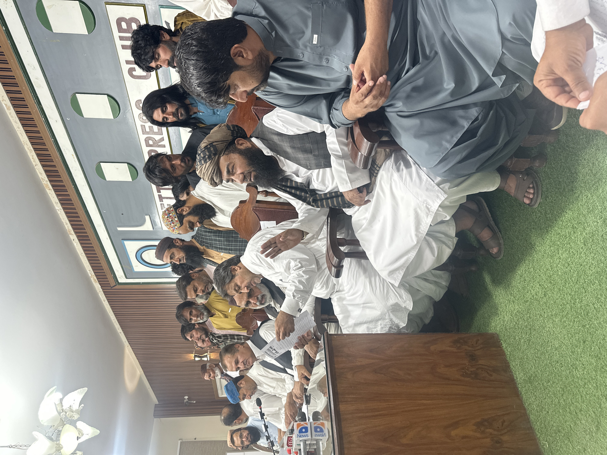 A group of men sit at tables in front of microphones in Quetta City Tehsil, Pakistan. Many of the men are dressed in traditional Pakistani clothing. The location appears to be a press club.