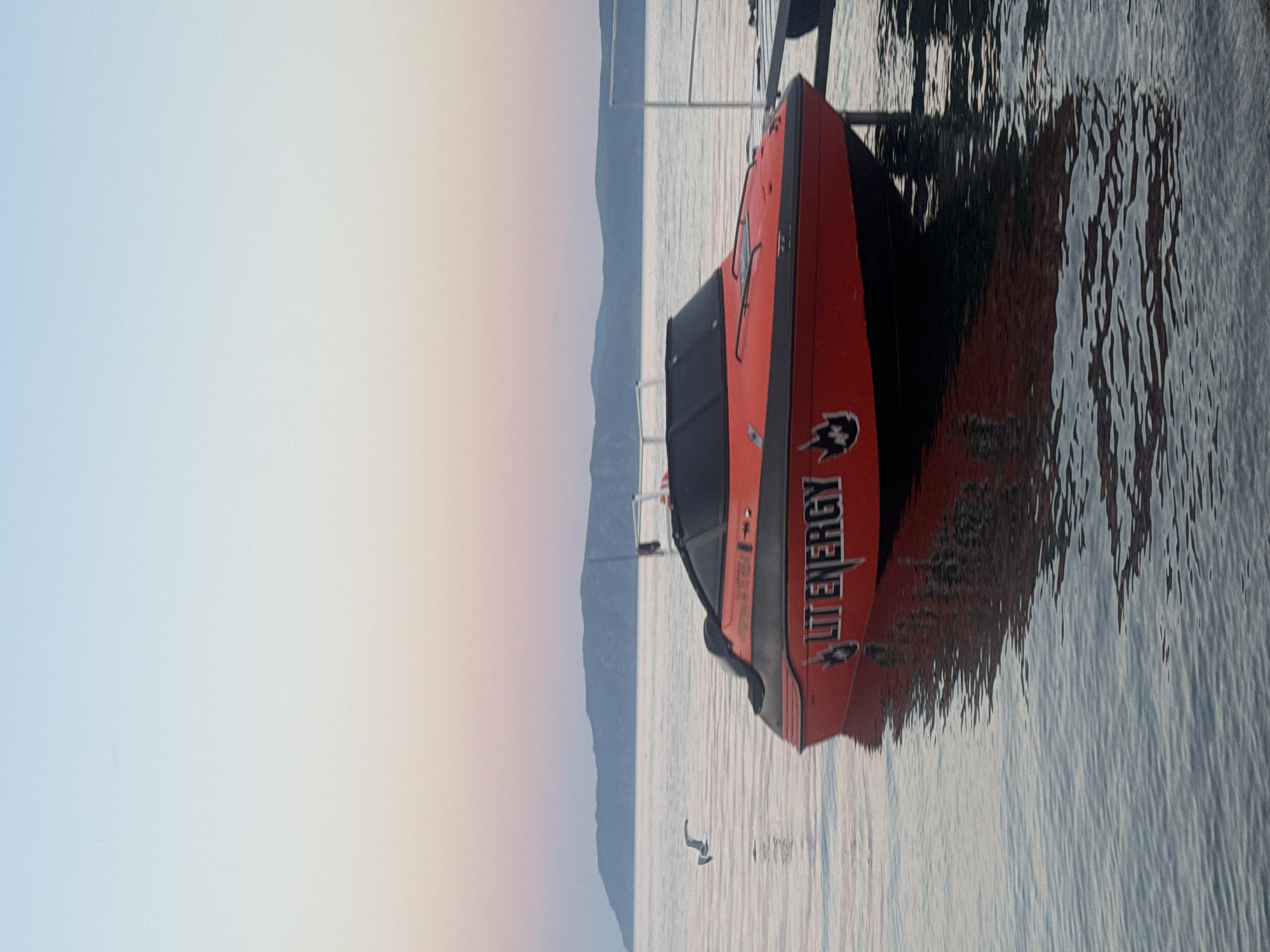 An orange and black speed boat with "LIT ENERGY" printed on its side is docked on calm water. The boat is positioned in the foreground, with its reflection visible on the water's surface. In the background, a hazy mountain range is visible against a soft, gradient sky transitioning from pale blue to a light pink hue. A white railing structure is also present to the right of the boat. A bird is in flight to the left of the boat, over the water. The scene appears to be a coastal or lakeside setting at dusk. The location is not the city of Listvyanka, Russia.