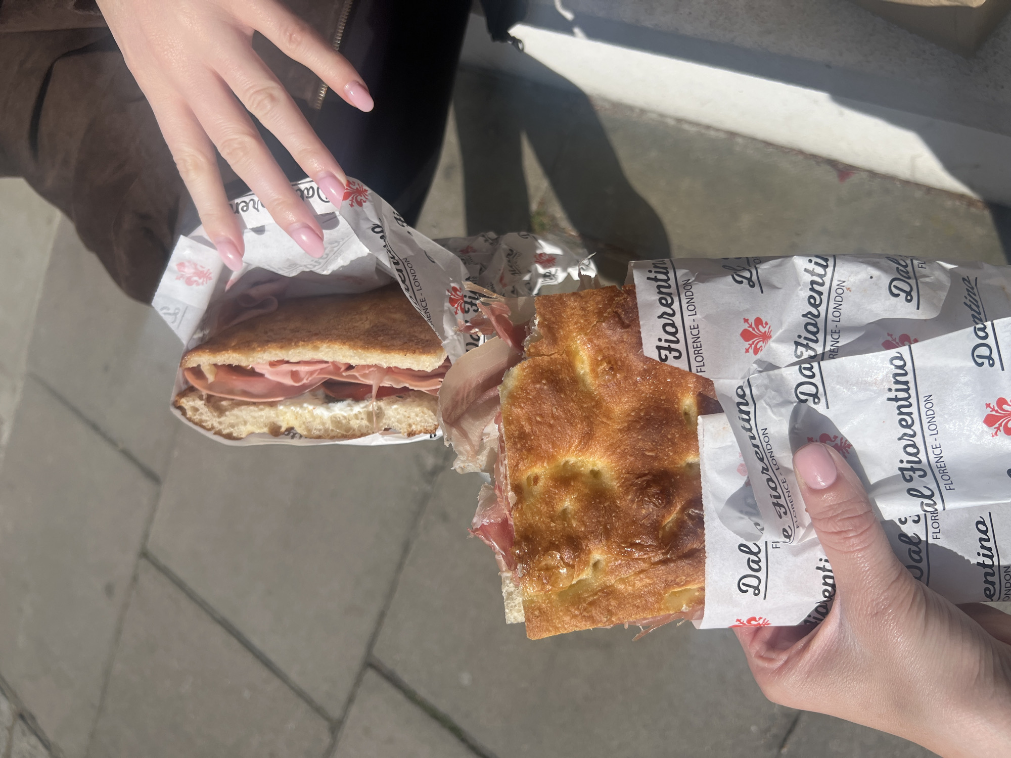 Two people are holding sandwiches wrapped in paper. The paper is white and says "Dal Fiorentino" and "Florence-London." One sandwich appears to be made with bread, meat, and cheese or a creamy sauce. The other sandwich is made with focaccia bread and meat. The people are standing on a gray stone or concrete surface. One person's hand is visible, with pale pink nail polish. The other person's hand is partially visible, also with pale pink nail polish. The image is taken outside, in sunlight.