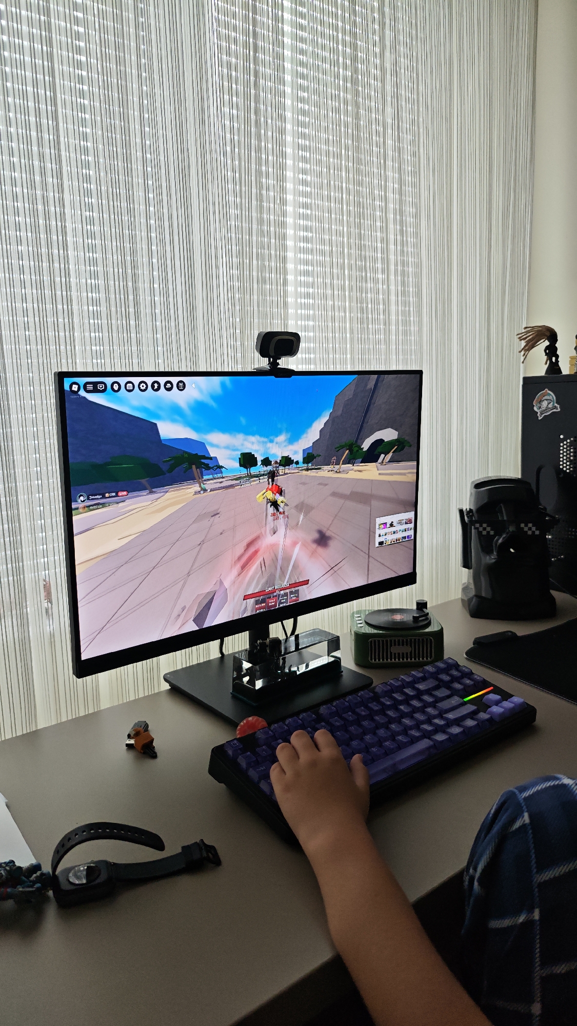 A person is using a purple mechanical keyboard in front of a computer monitor displaying a Roblox game, likely taken indoors in a home or office environment. The monitor has a webcam attached on top, and several items are placed on the desk: a smartwatch, a Lego figure, a candy, and a miniature record player. A Star Wars-themed Darth Vader figurine with sunglasses, and a computer tower are visible in the background on the right side of the desk. The window behind the monitor is covered with white string curtains. The person's arm and part of a plaid-patterned shirt or blanket are visible. The location is not identifiable.