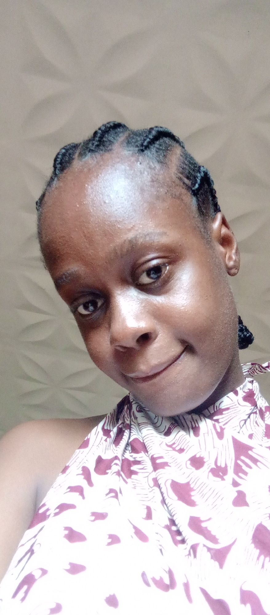 A close-up portrait features a young woman, likely in Akwanga, Nigeria, looking directly at the camera with a gentle, closed-mouth smile. She has dark skin with a visible natural sheen on her forehead, nose, and cheeks. Her dark hair is styled in neat, tightly braided cornrows that frame her face. She is wearing a halter-neck top with a white base patterned with abstract, reddish-brown or maroon organic shapes. The background consists of a light-colored wall with a subtle, repeating three-dimensional pattern, possibly floral or star-like, visible behind her head and to her left. The lighting is soft and even, suggesting an indoor setting during the daytime. No text is visible in the image.